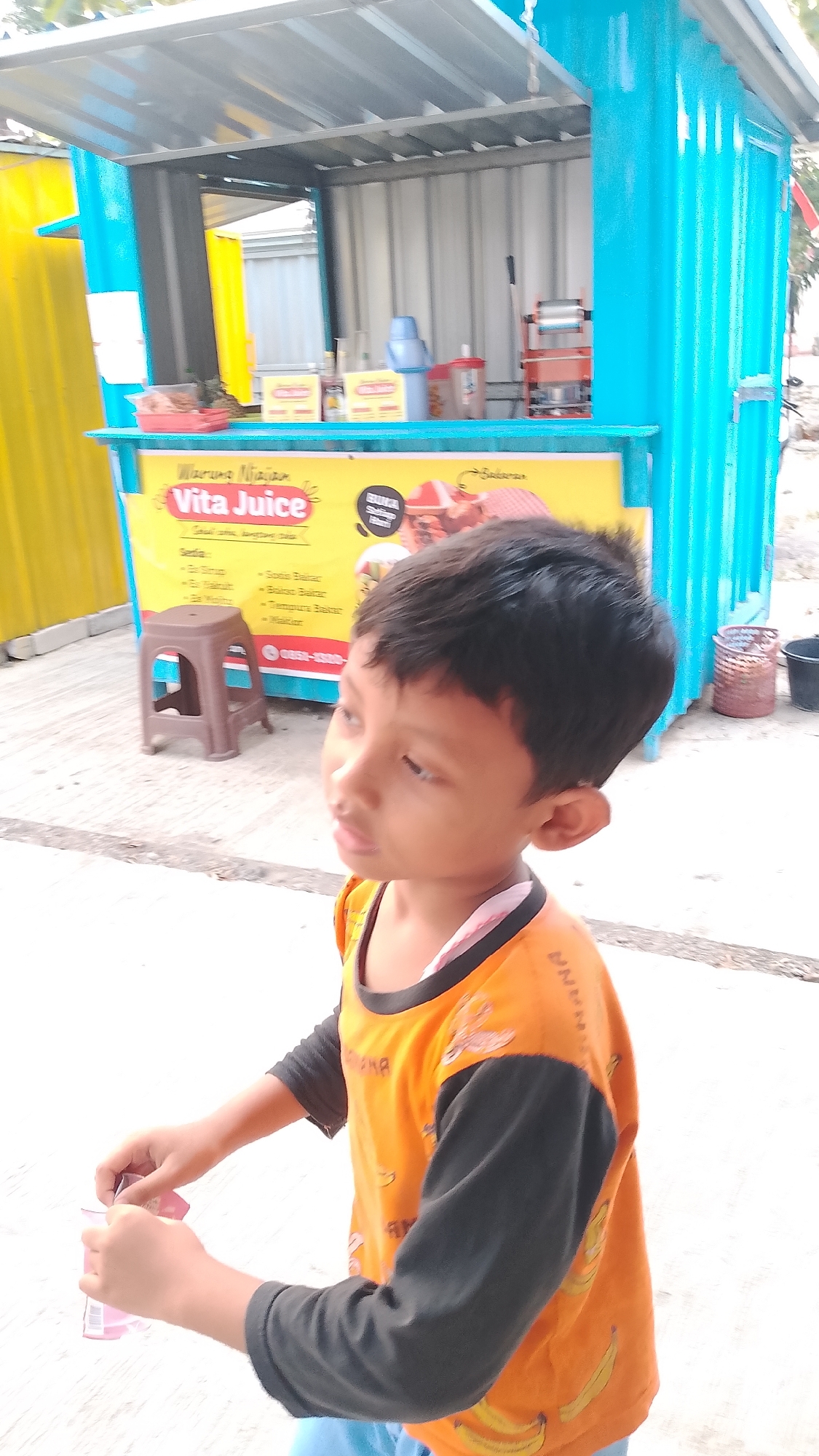 A young Indonesian boy, appearing to be around five to seven years old, stands in front of a brightly colored food cart in Karanglangu, Indonesia. The cart is painted a vibrant teal and yellow, with "Vita Juice" prominently displayed on a yellow banner. The boy wears an orange long-sleeved shirt with a banana print and blue shorts. He is holding a small pink packet in his hands and looking off to the side. The cart is stocked with various drinks and snacks, and a small brown stool sits to the left of the cart. The scene appears to be outdoors, with a paved ground and a corrugated metal roof over the cart. The overall impression is of a typical street scene in a local Indonesian town.