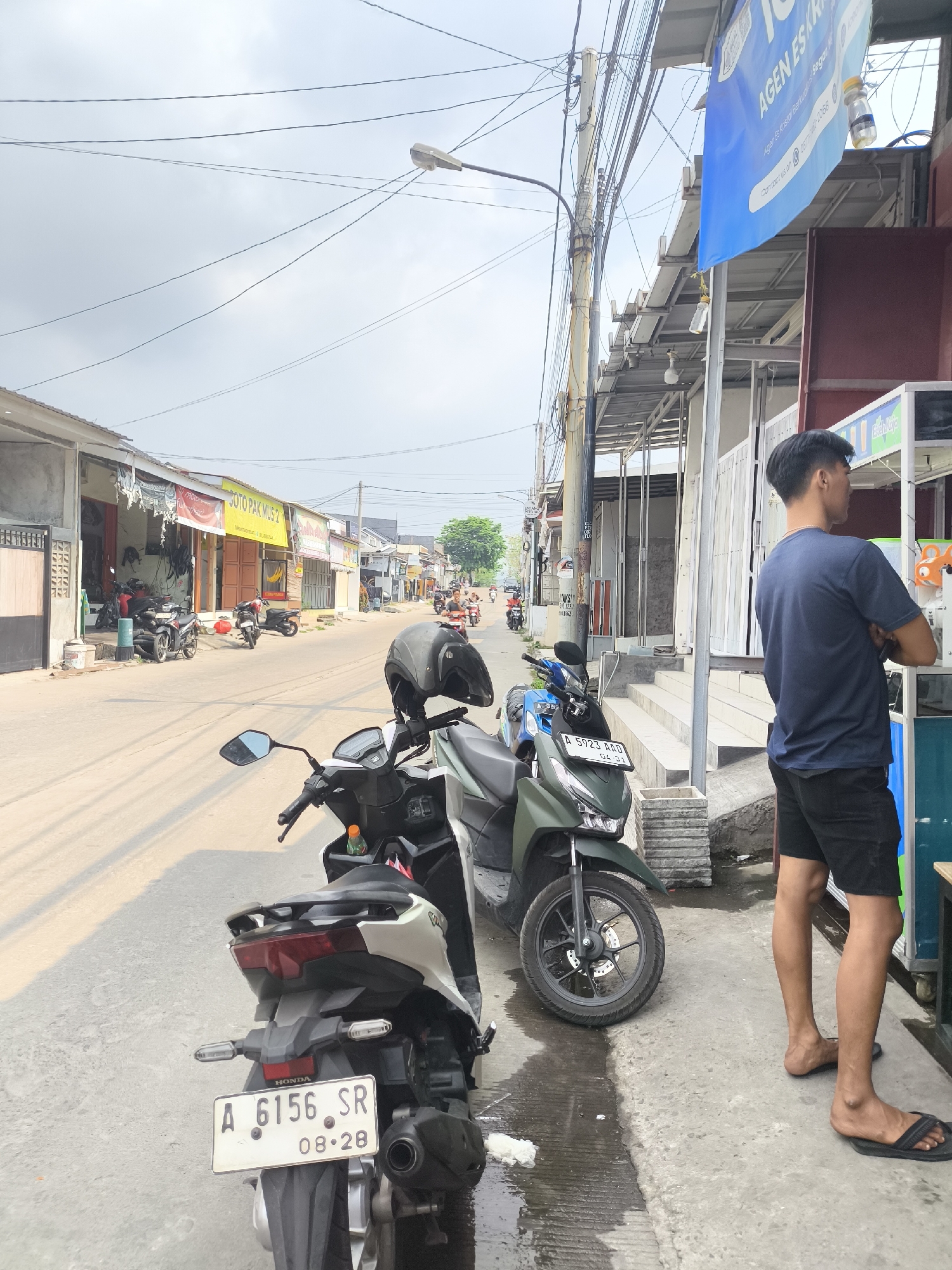 The image captures a street scene in Cilegon, Indonesia, likely a commercial or residential area with a row of buildings lining one side of the road. In the foreground, several motorcycles are parked along the roadside, with one white and grey scooter prominently displayed, featuring a license plate that reads "A 6156 SR 08-28". Another dark green scooter is parked next to it. A man wearing a dark blue t-shirt and black shorts stands facing away from the camera, looking towards a food or drink stall, and wearing black flip-flops. A helmet rests on the handlebars of the white and grey scooter. The street appears to be paved, and there are overhead electrical wires crisscrossing the sky. The buildings on the left side of the street have awnings and display signs, one of which indicates "SOTO PAKMUS 2". The overall environment suggests a casual, everyday setting in a Southeast Asian city.