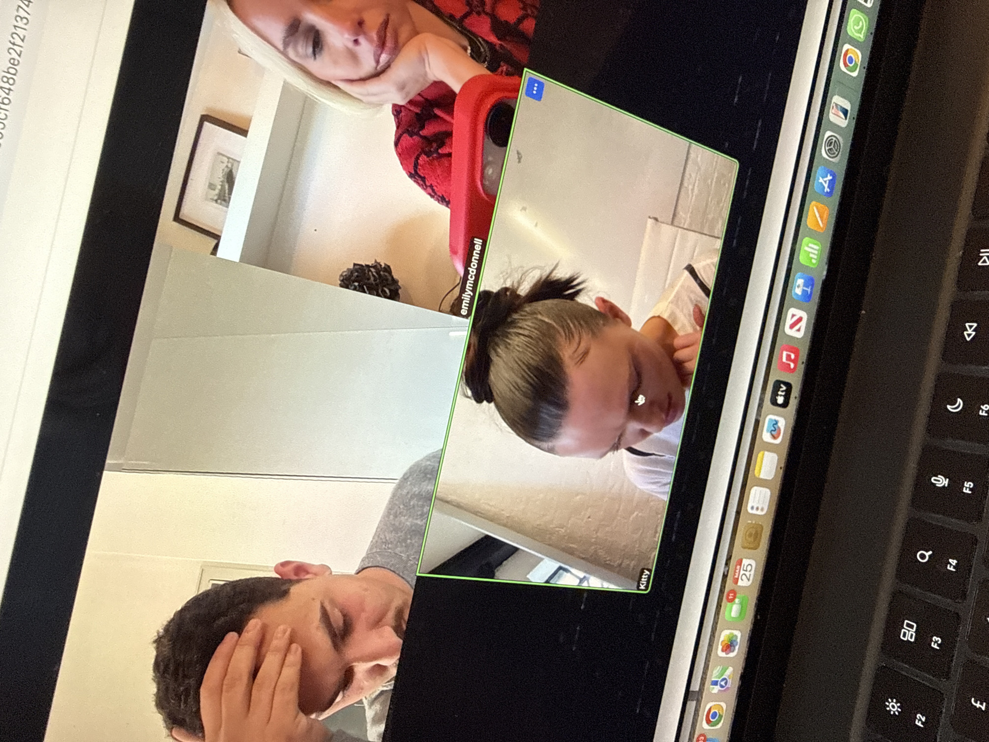The image displays a laptop screen captured from an overhead perspective, depicting a video conference call involving three participants, with a portion of the laptop's keyboard visible at the bottom.
In the upper left quadrant of the screen, a male participant with dark, curly hair and stubble is shown wearing a grey shirt, with his right hand resting on his forehead and his gaze directed downwards. In the upper right quadrant, a female participant identified as "emilymcdonnell" is visible, characterized by blonde hair and a red and black patterned top. She rests her head on her left hand, looking downwards. A red-cased mobile phone is positioned in front of her. Her background features a white shelving unit on which a framed, monochrome print is displayed. This print clearly depicts a cityscape, specifically identifiable as a view of the Houses of Parliament and Big Ben from across the River Thames, indicating a location within the city of London, United Kingdom. In the lower quadrant, a female participant named "Kitty" is shown, with dark hair pulled into a high bun, her head tilted downwards. A white mouse cursor is visible near her forehead on the screen.
The lower portion of the image reveals the laptop's keyboard, displaying function keys F2 through F6, which include symbols for screen brightness, a search function, a microphone, and a crescent moon. The '£' (pound sterling) symbol is visible on a key. The macOS Dock is present at the bottom of the screen, displaying icons for various applications such as Google Chrome, Calendar (showing "MAR 25"), Apple TV, and Music.