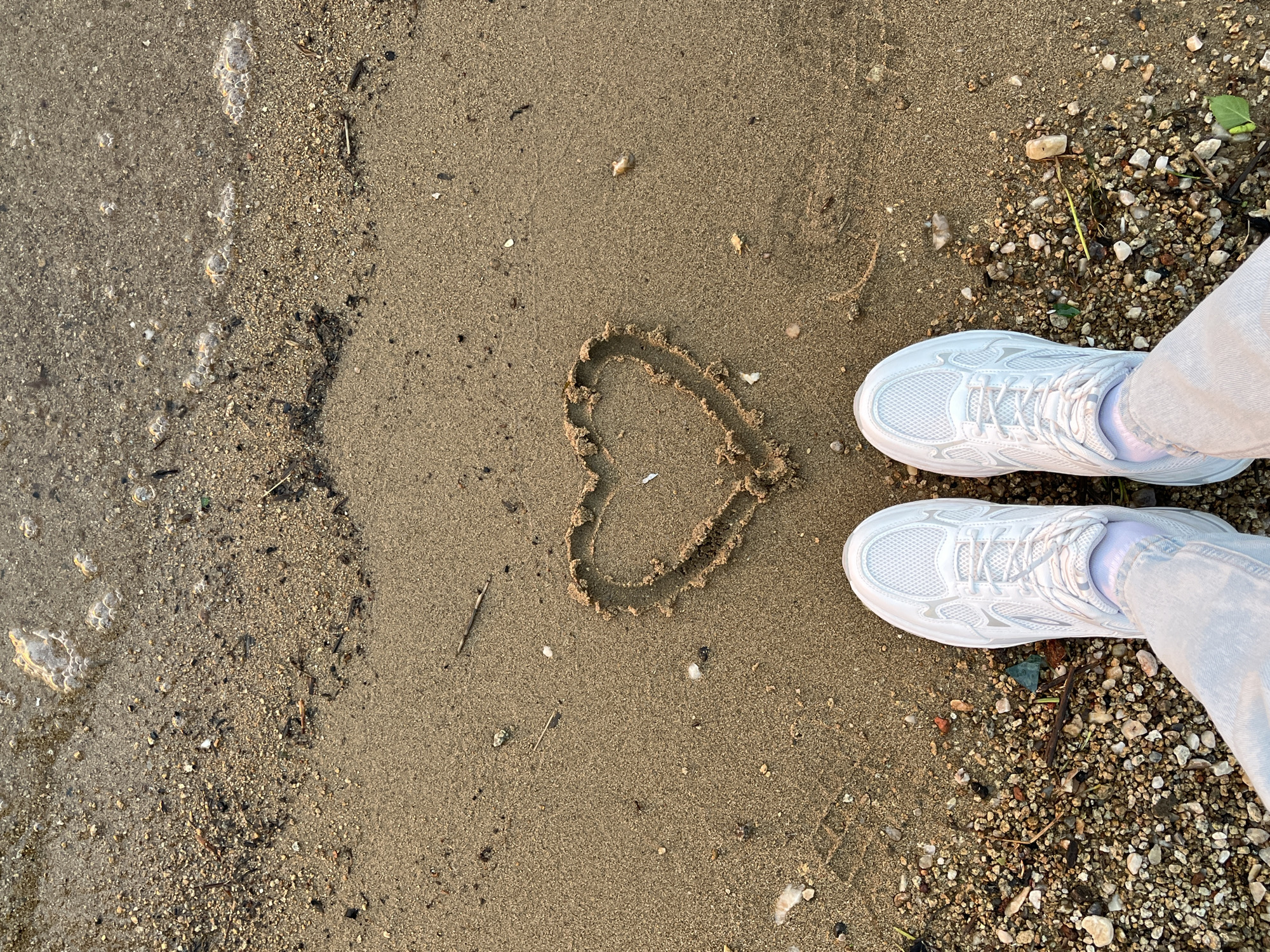 The image shows a beach scene. A heart is drawn in the sand. Two white sneakers are situated to the right of the heart, partially on a section of the beach with small rocks. The sand is wet near the left side of the frame. It is not possible to determine the specific location of the beach in Yekaterinburg, Russia from this single image.