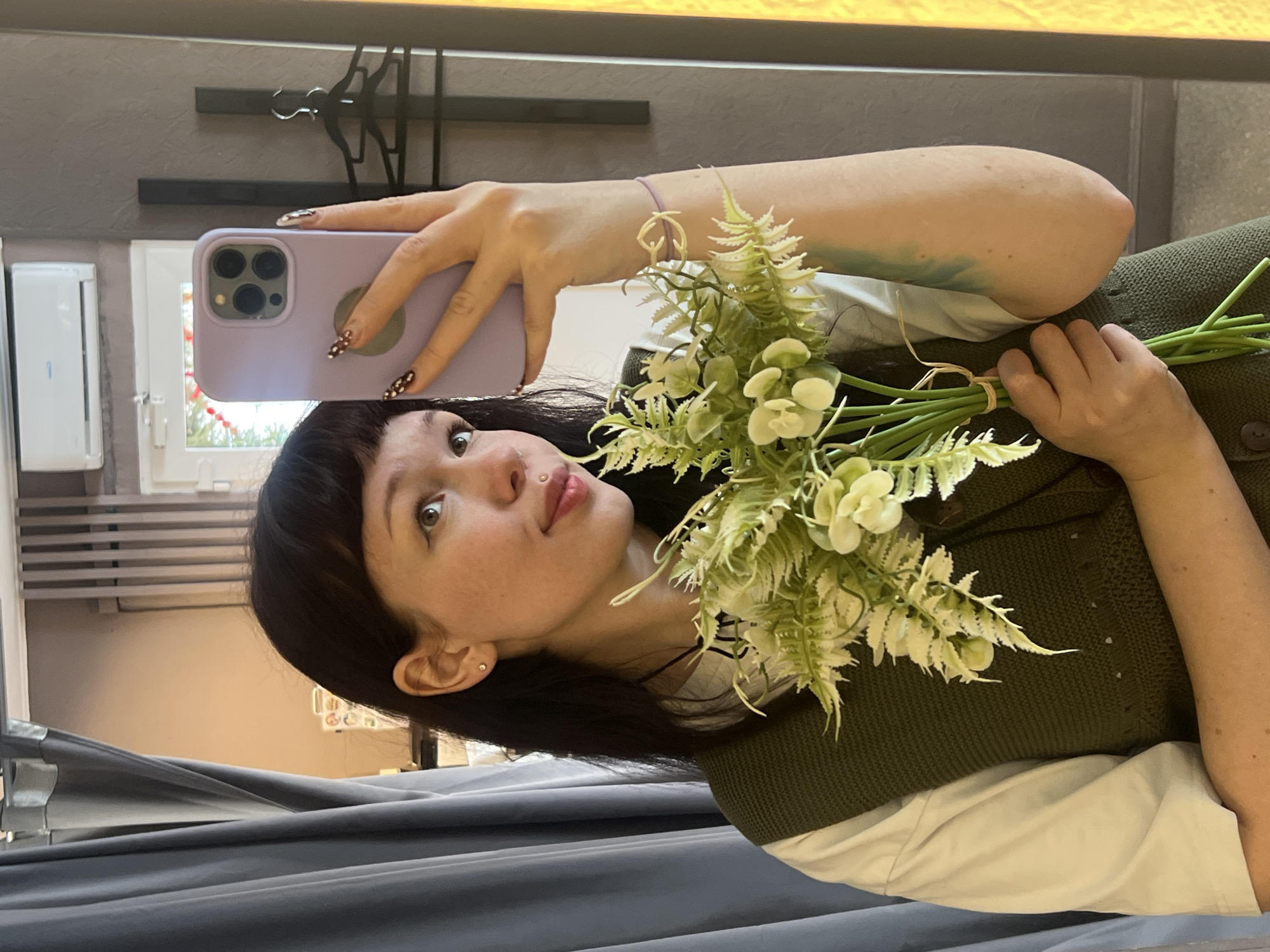 A young adult female is depicted in a mirror selfie taken in Chaikovsky, Russia. She has dark hair with bangs, light skin, a visible lip piercing above the upper lip, and an earring in her left earlobe. She is wearing a white shirt underneath a dark green knitted vest. In her left arm, she holds a bouquet of artificial green and white flowers tied with twine. Her right hand, featuring nails with a dark patterned design, holds a light purple smartphone, visible with its three camera lenses, used to capture the image. A light purple bracelet with a gold-colored heart-shaped charm is on her right wrist, and a faint light blue tattoo is visible on her inner right forearm. The woman's gaze is directed slightly upwards and to her right. The background, reflected in the mirror, includes grey curtains on the left, a white air conditioning unit mounted high on a wall, a window partially obscured with red lights, and dark vertical paneling with a black coat hanger on the right.