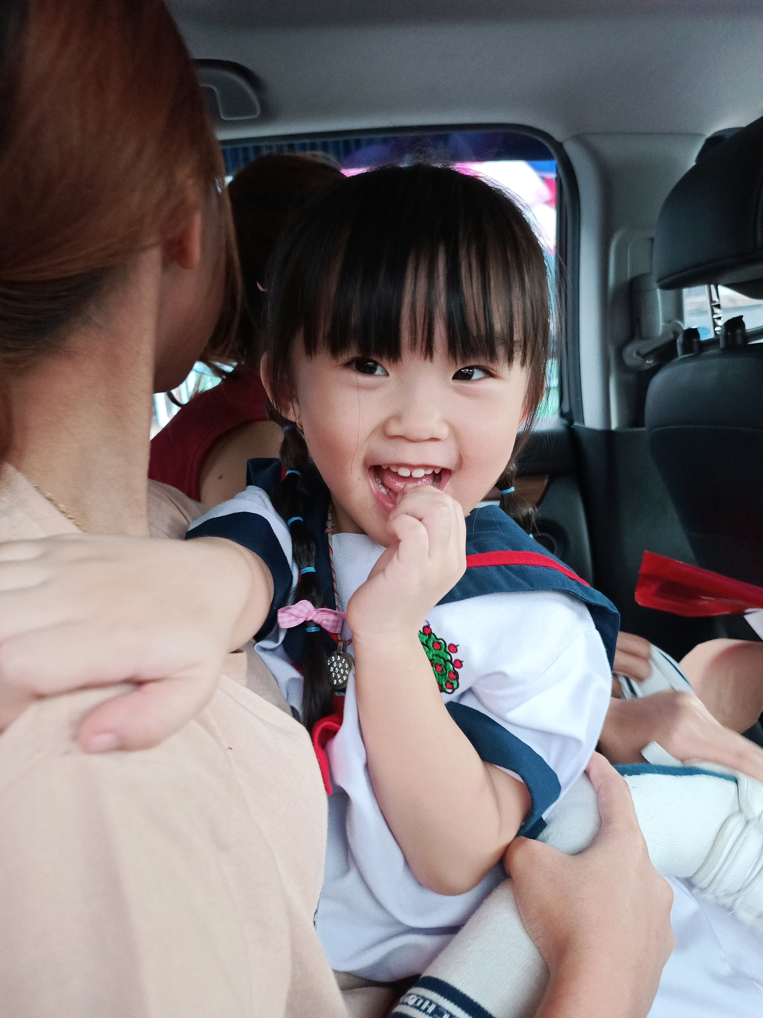In Pakulonan Barat, Indonesia, a young East Asian girl with black hair in pigtails, adorned with pink ribbons and blue ties, smiles widely in a car. She is wearing a white school uniform with navy trim and a red collar, featuring a green embroidered emblem on the left chest. Her right hand is raised to her mouth. She is seated next to an adult, whose shoulder and back are visible in the foreground, partially obscuring the girl. The background shows the interior of a car, including a black car seat, a window with a blurred view of the outside, and possibly another person behind the girl. The lighting suggests an indoor setting, likely during the day.
