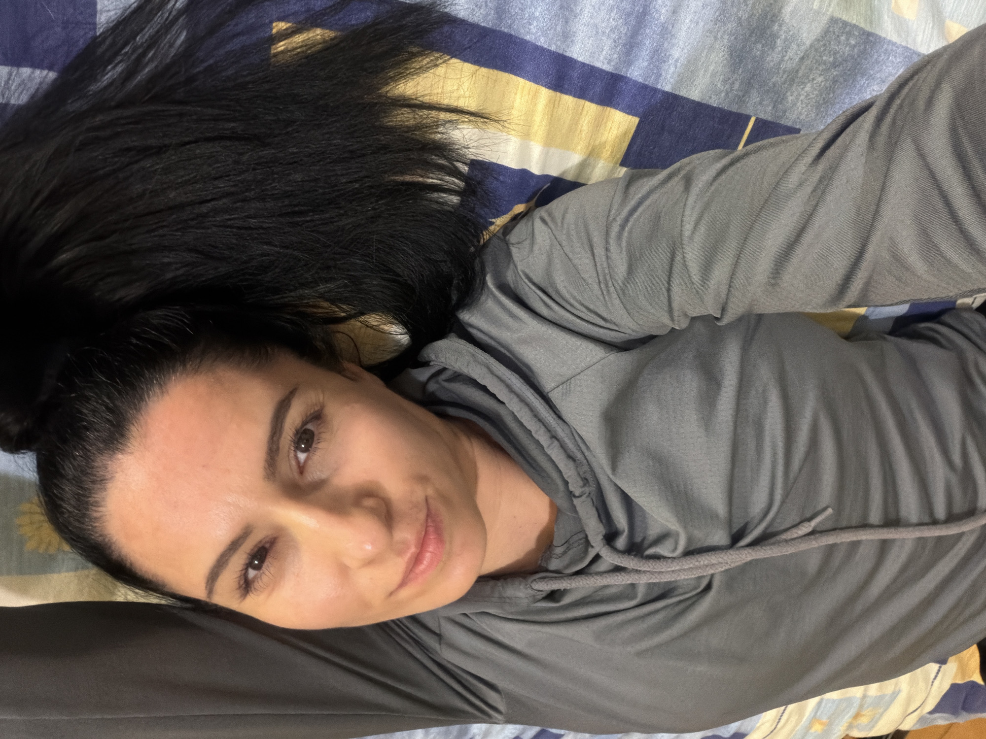 A single individual is depicted lying horizontally on a patterned bedsheet within an indoor setting in Pleven, Bulgaria. The subject, an adult female, possesses dark hair, dark eyes, and a light complexion. She is dressed in a grey long-sleeved top, featuring a visible grey drawstring around the neckline. Her head is tilted slightly towards the camera, with her gaze directed forward, exhibiting a slight smile. Her dark hair is spread across the bedsheet to her right. The bedsheet displays a repeating pattern composed of blue, white, and golden/yellow rectangular or striped elements. The illumination is soft and even.