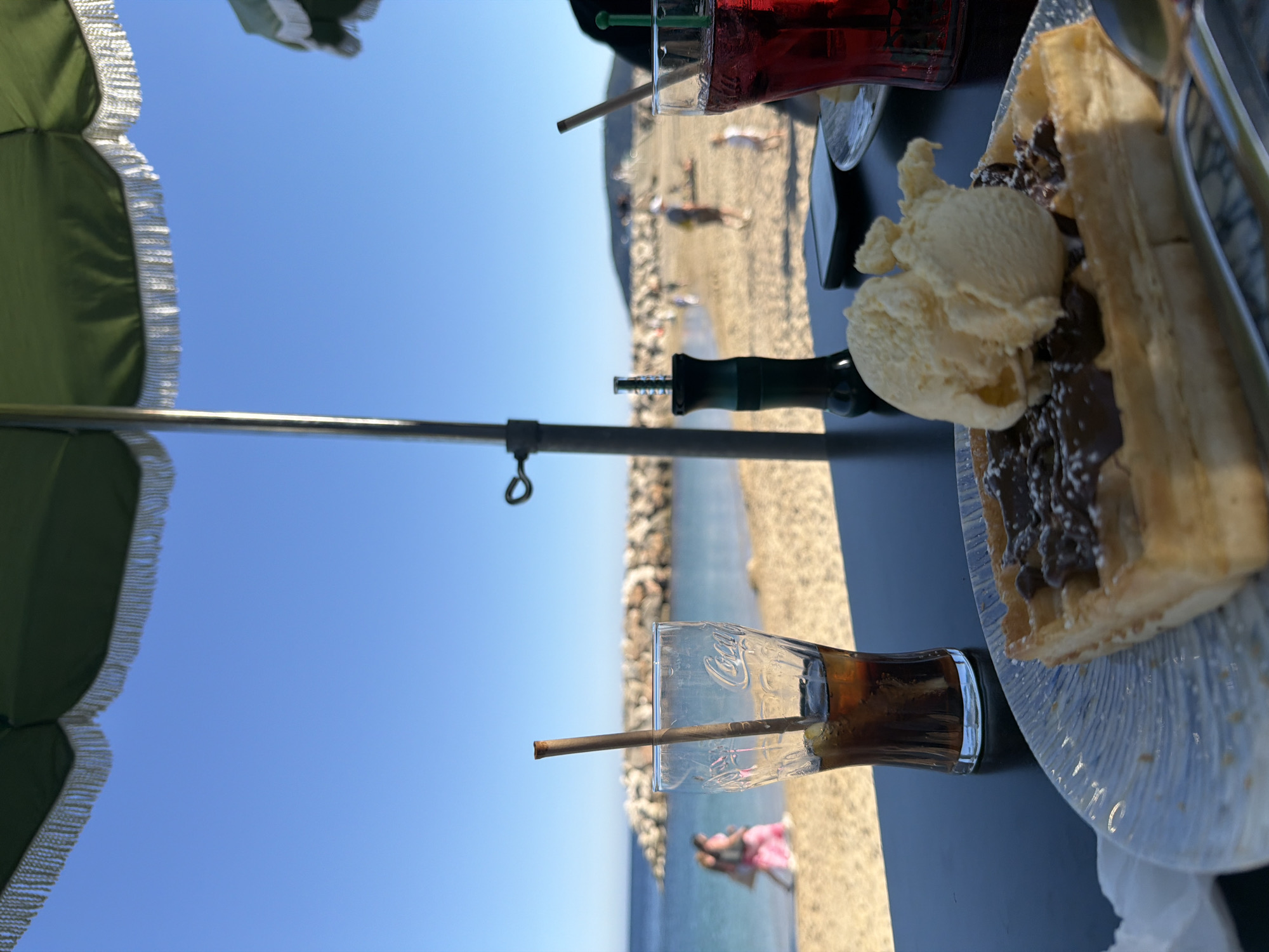 The image depicts a vibrant, sunny scene at a beachfront cafe, likely in Toulon, France. In the immediate foreground, a dark table holds a delicious dessert and beverages. A golden-brown waffle, generously covered in melted chocolate sauce and sprinkled with what appears to be powdered sugar or nuts, is presented on a clear, textured plate. Alongside it are two scoops of creamy vanilla ice cream.
To the right of the waffle, a clear glass displays a visible "Coca-Cola" logo, containing a dark, fizzy beverage with a straw. Another glass, partially visible at the top, holds a bright red drink with a green straw. A dark green hookah or shisha pipe is also on the table, contributing to the relaxed atmosphere.
A large, dark green umbrella with decorative white fringing is partially visible on the left side of the frame, providing shade and confirming an outdoor seating arrangement. The background features a wide, sandy beach where several blurry figures of people can be seen, enjoying the sun and shore. Beyond the beach, the calm, clear blue sea extends to the horizon, dotted with a rocky breakwater. The sky above is a brilliant, cloudless blue, indicating fine weather. The scene exudes a sense of leisure and enjoyment by the coast on a bright day.