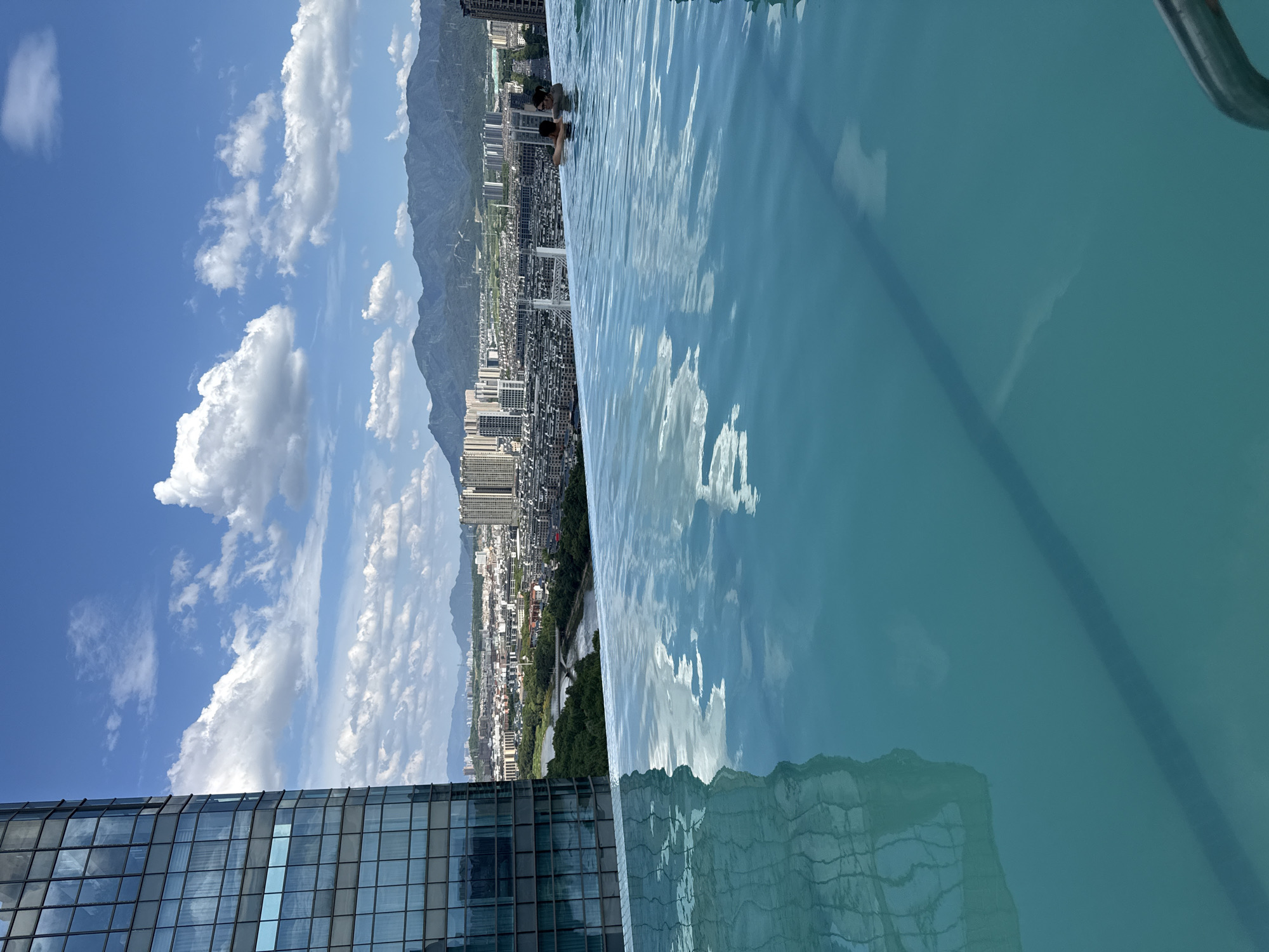 This is an eye-level, vertical, medium shot of an infinity pool overlooking a cityscape. The pool is in the foreground, its surface reflecting the sky and surrounding buildings. Two individuals are partially submerged in the water in the middle ground. In the background, a dense urban area with numerous buildings and a river is visible, with mountains forming the horizon. A tall glass-fronted building dominates the left side of the frame. The sky is blue with scattered white clouds. The image was taken in Yiwu, China.