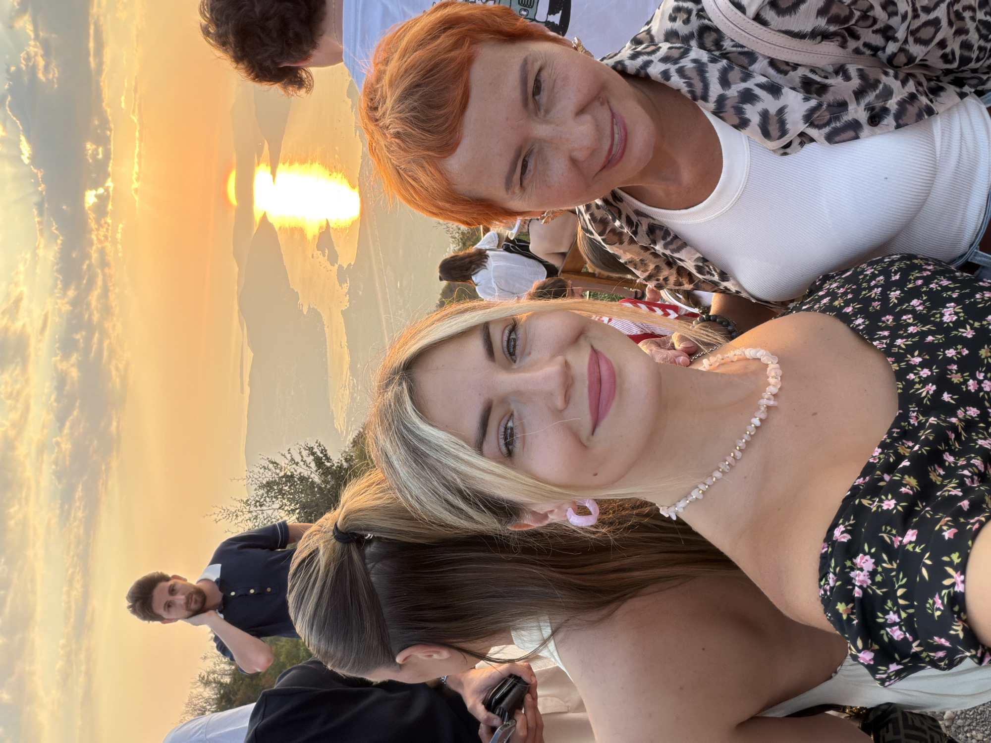 The image captures a group of people at a scenic overlook in Kuk, Montenegro, during sunset. The sun is setting over a body of water, casting a bright reflection. In the foreground, two women are close to the camera. The woman on the right has short, vibrant orange hair and is wearing a white tank top under a leopard-print jacket. She is smiling. The woman on the left has blonde hair and is wearing a floral off-the-shoulder top and a light pink beaded necklace. She is also smiling. Behind them, partially visible, are other individuals. A man with dark hair is standing in the background to the left, wearing a dark blue polo shirt. Further back, another person in a white shirt is visible. The overall scene suggests a social gathering enjoying the natural beauty of the location.