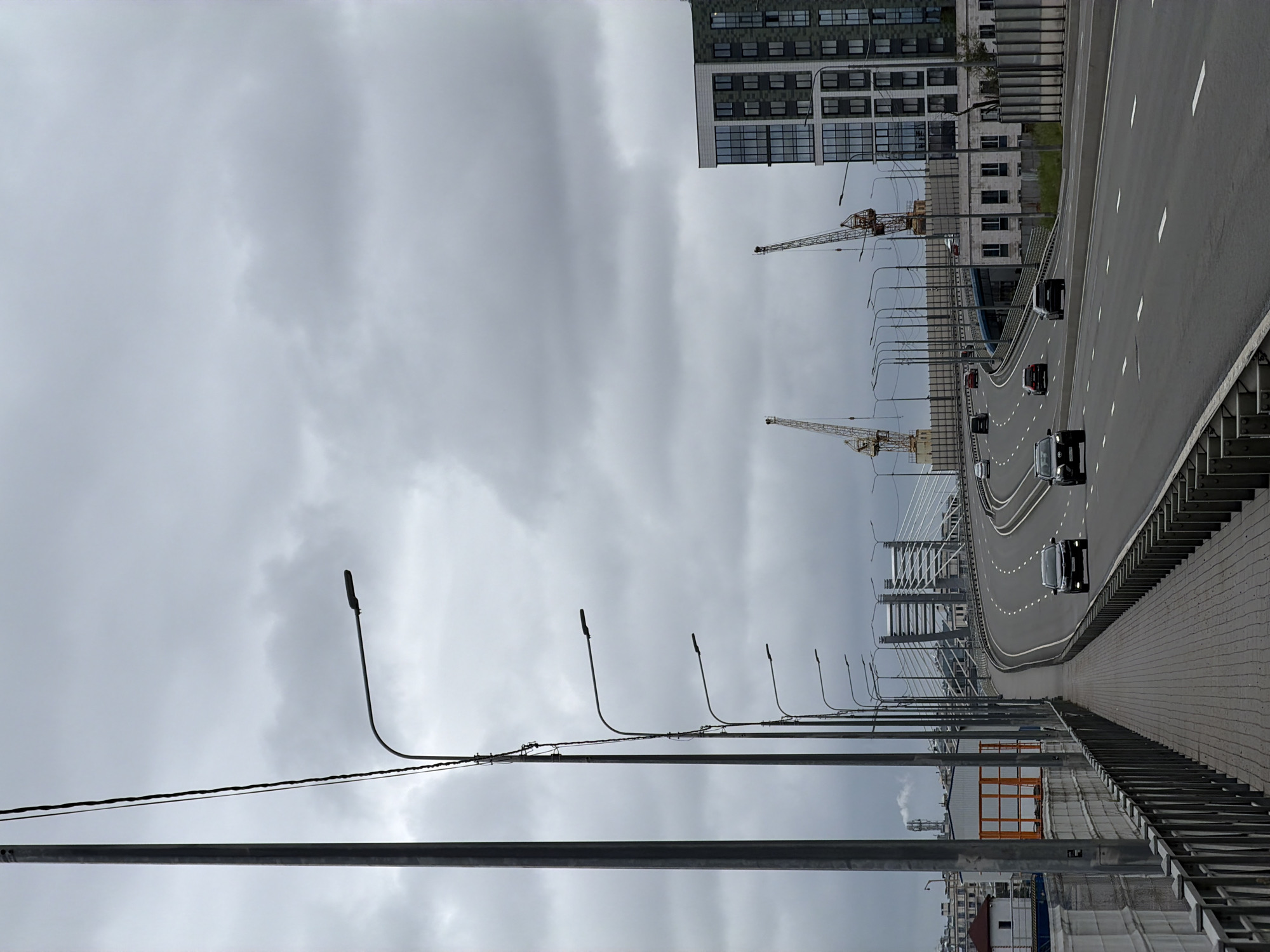 The image depicts a view of an overpass or elevated highway in Saint Petersburg, Russia, under a cloudy sky. On the left, a tall lamppost stands prominently, accompanied by a series of smaller lampposts lining the walkway. The walkway itself is paved with grey bricks and bordered by a metal fence. The highway has multiple lanes and is occupied by several cars driving in the same direction.
In the background, industrial structures, including cranes, are visible, suggesting ongoing construction or proximity to a port or industrial area. A modern, multi-story building with a glass facade is situated on the right side of the image. The overall atmosphere is overcast and somewhat industrial.