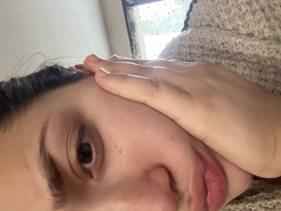 A close-up image captures the left side of a female's face and upper torso, taken from a slightly upward angle, consistent with a selfie. The subject possesses medium skin tone, a dark brown eye, dark hair pulled back, and full, slightly pursed lips. Her left hand is positioned with fingers resting against her left cheek and palm against her jawline. She wears a beige or light-colored knitted sweater. The background, out of focus, reveals a window letting in bright natural light, partially covered by a translucent material or curtain featuring a pattern of small, scattered multicolored circles in shades of red, orange, green, and blue. The scene is indoors. Visual evidence within the image does not permit confirmation of the specified location in Ancenis, France.