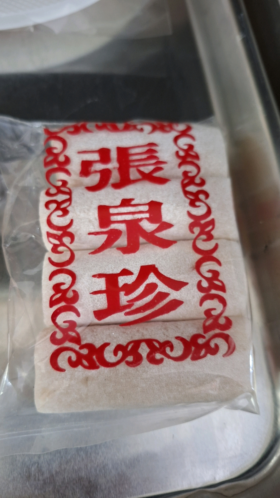 A close-up, vertical shot shows a package of white, cylindrical snacks sealed in clear plastic. Red Chinese characters are printed on the packaging, arranged vertically. The snack appears to be layered, with at least three distinct rolls visible. The plastic packaging is slightly crinkled. The background is blurred, but suggests a metallic surface, possibly a tray or shelf, and a hint of white material at the top. The lighting is soft, casting gentle shadows.
