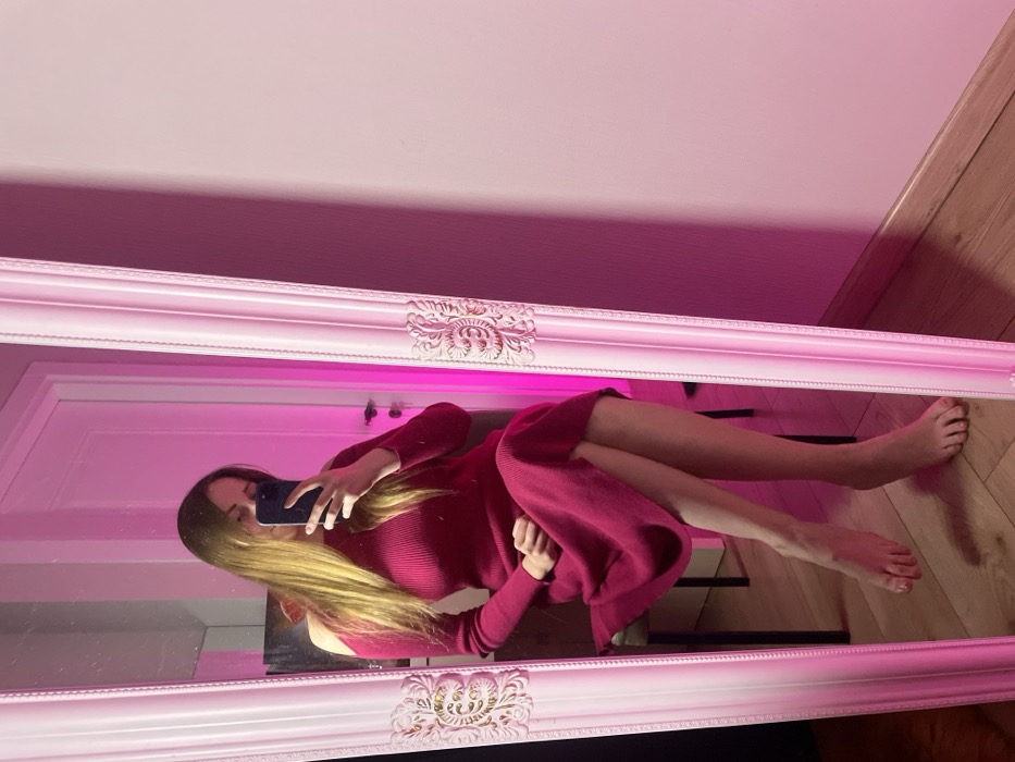 An indoor scene in what appears to be a private room within the city of Batumi, Georgia. The specific address or distinguishing landmarks within Batumi are not discernible. The image captures a female subject reflected in a full-length mirror. She has long, light brown hair and is seated on a chair, holding a dark-colored smartphone to take a mirror selfie, which obscures her upper face. She is wearing a fuchsia-colored, ribbed, long-sleeved midi dress featuring cut-out shoulders. Her legs are crossed, and she is barefoot.
The mirror possesses a white, ornate frame with decorative carvings. The room is illuminated by a strong pink ambient light source, which casts a magenta hue on the plain wall visible to the right of the mirror and imparts a pink glow to the mirror's frame. The floor is constructed of light-colored wooden planks, visible at the bottom of the frame, meeting a light-colored baseboard. In the mirror's reflection, behind the subject, a white door with a silver handle is visible, along with a partial view of a dark object or artwork on the wall above the chair.