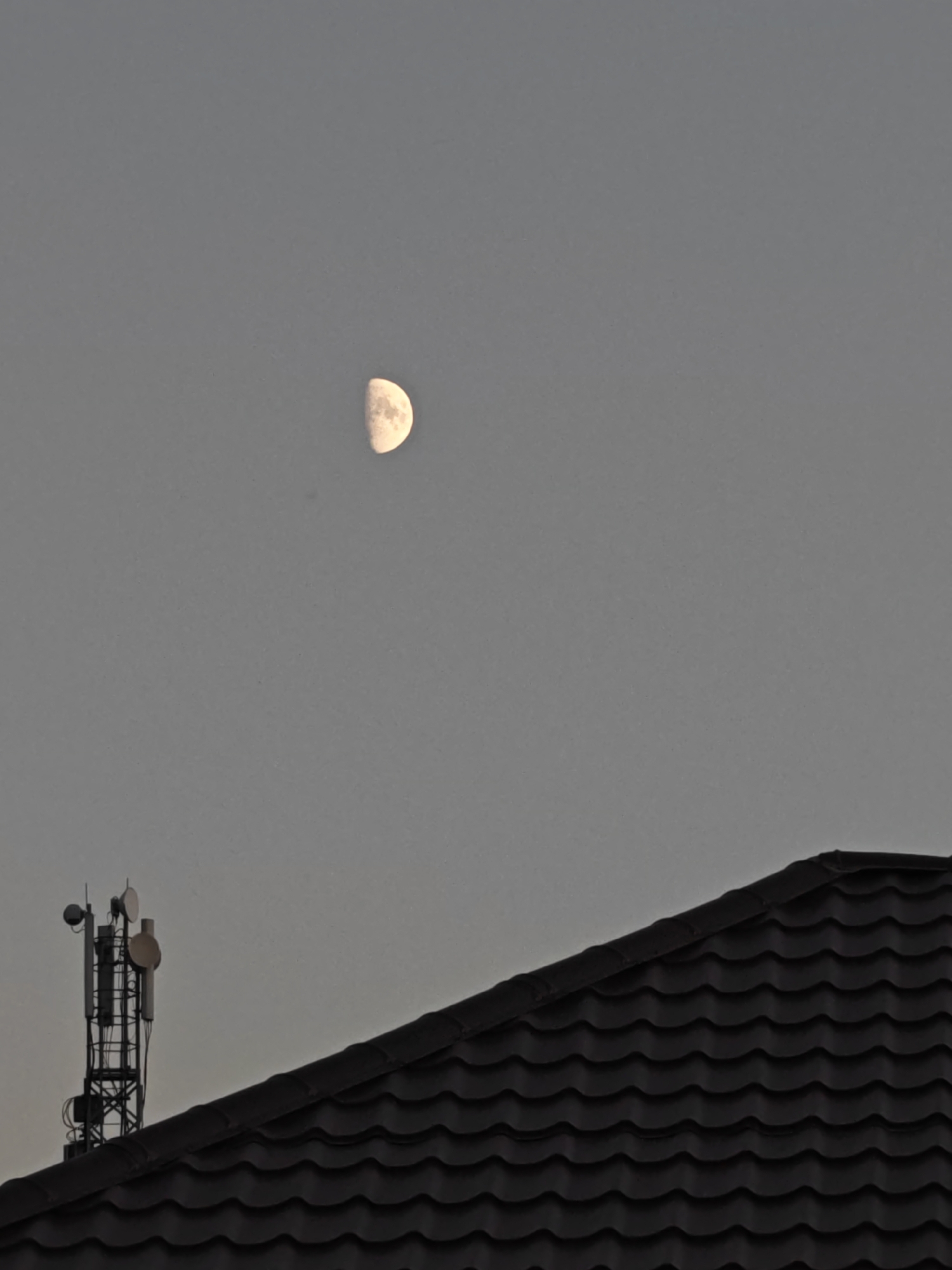 The image shows a cloudy sky with a crescent moon visible in the upper left quadrant. In the lower portion of the image, the dark, tiled roof of a building occupies the foreground. To the left of the roof, a tall, metal telecommunications tower with multiple antennas is visible against the sky. The scene appears to be set in Vodopadnyy, Russia.
