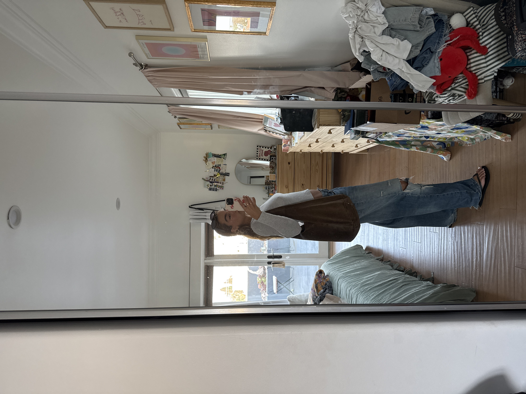 A young woman is captured in a mirror selfie, standing upright in what appears to be a bedroom or dressing area. She is holding a smartphone, with a distinct patterned case, up to take the picture of her reflection.
She is dressed in a light grey, long-sleeved top, distressed blue jeans with rips on the knees, and black flip-flops. A brown shoulder bag is visible on her side, and sunglasses are perched on her head.
The reflected interior shows a light-colored wooden dresser with multiple drawers, topped with various items, including a small mirror and an assortment of decorative objects. Above the dresser, framed artwork hangs on a light-colored wall. One frame contains handwritten text that includes "Harley's got your back" and "XOXO". Pink curtains partially obscure a window or glass door. To the right, a large pile of clothes, including a red plush toy (resembling a crab or octopus) and striped fabric, is stacked on a surface.
Through the reflected window or door, a bright outdoor scene is visible, suggesting daytime with good natural light. The exterior view includes what appears to be a patio or backyard with a white outdoor chair and some greenery. A bed with a light green ruffled duvet is visible in the lower part of the reflection. The room has light wooden flooring. The entire image is rotated 90 degrees counter-clockwise.