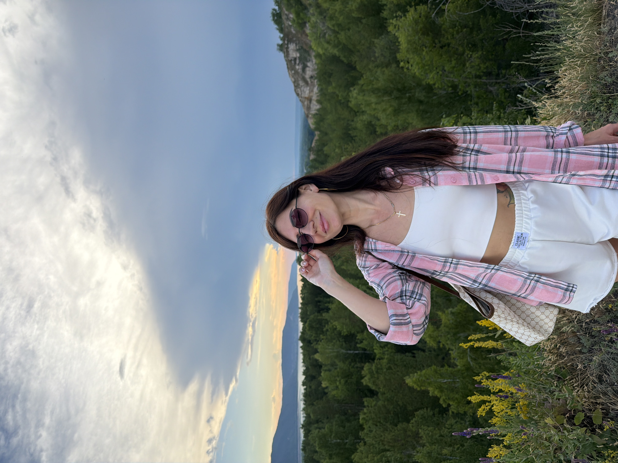 A woman stands on a hillside overlooking a forest and a body of water. She has long, dark hair and is wearing sunglasses, a pink and white plaid shirt, a white crop top, and white shorts. She is holding her sunglasses and smiling. The background shows a dense green forest, a wide river or lake, and hills in the distance. The sky is cloudy with patches of sunlight. The location is the city of Volzhsky, Russia, near the Volga River.