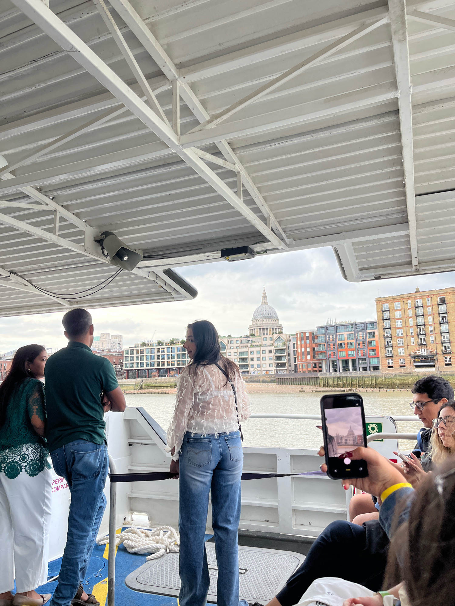 This is a medium shot, taken from a boat on the River Thames, looking towards the London skyline. In the foreground, a person's hand holds up a smartphone, taking a picture of St. Paul's Cathedral and the surrounding buildings. The cathedral, with its distinctive dome, is a prominent landmark. The boat's white metal ceiling with exposed beams is visible at the top of the frame. People are standing and sitting on the boat. To the left, a woman in a green top and white pants stands with her back to the camera, looking out at the view. Next to her is a man in a green polo shirt and jeans, also facing away. To the right of the man, a woman in a white lacy top and blue jeans stands. Further to the right, a man wearing glasses is visible, and another woman is partially visible in the lower right corner. The river is calm, and the sky is overcast. Buildings with modern and traditional architecture line the riverbank.