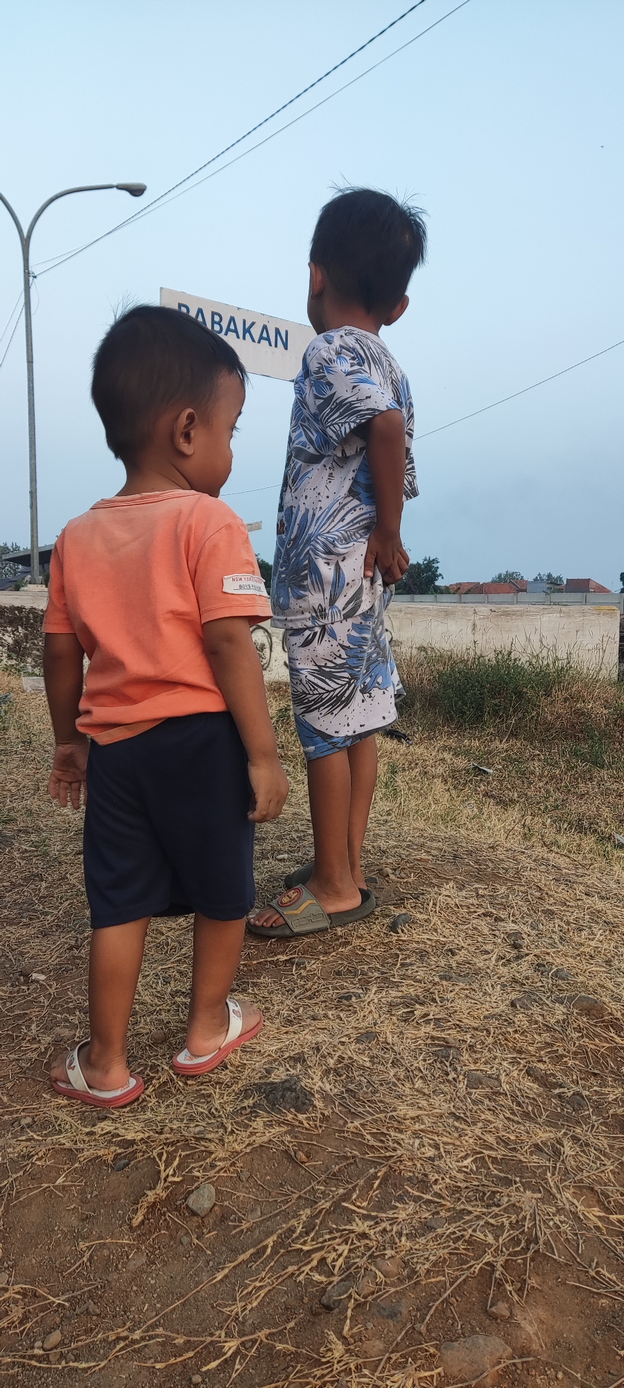 Two young boys stand outdoors on a dry, grassy area. The boy on the left faces away from the camera, wearing an orange t-shirt and navy shorts. He has dark hair and is wearing light-colored sandals. The boy on the right also faces away from the camera, wearing a white t-shirt with a blue floral pattern and matching shorts. He has dark hair and is wearing dark-colored slides. Behind them, a white sign with blue text that reads "BABAKAN" is visible, along with a street lamp and power lines against a hazy sky. The ground is covered in dry grass and dirt, with some scattered rocks. In the background, there is a low wall and some buildings with red roofs. The scene appears to be a street or roadside location in Pakusamben, Indonesia.