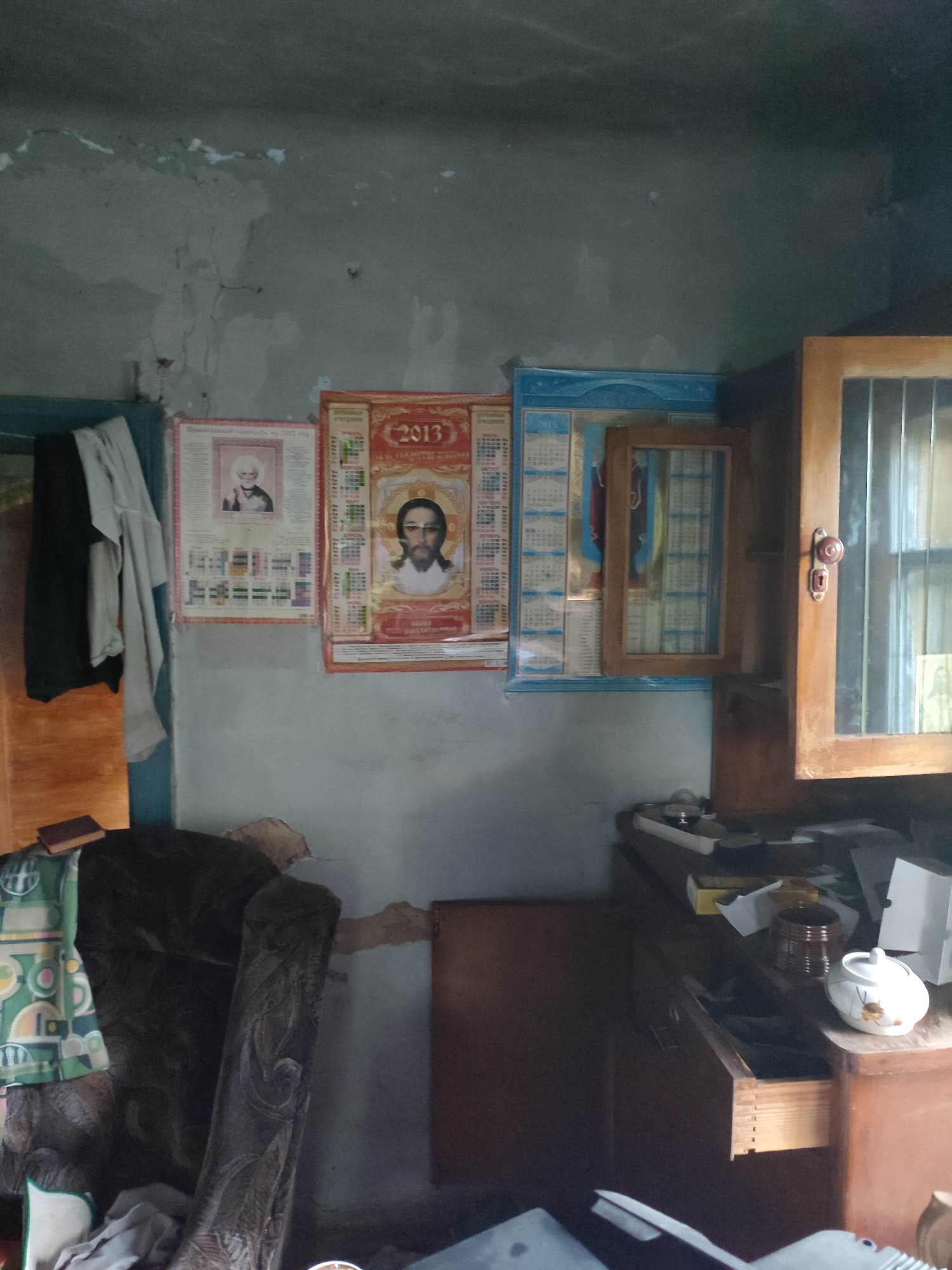 The image depicts a cluttered, dimly lit interior that appears to be a room in a residential dwelling in Gremyachinsk, Russia. The walls are in poor condition, with visible cracks and peeling paint. On the wall, there are three calendars hanging. The leftmost calendar features a portrait of an older man with white hair and a beard. The middle calendar has a portrait of a man with dark hair and a beard, and prominently displays the year "2013". The third calendar, framed in blue, also shows a grid of dates and appears to have a small wooden framed glass door slightly ajar on its front.
To the left, a dark-colored armchair with intricate carvings is visible, partially covered by some fabric. Next to it, a wooden cabinet with glass doors stands open, revealing items stored inside. On a wooden desk or counter below the cabinet, several objects are scattered, including a small white teapot with floral accents, a brown cylindrical container, some paper, and what looks like a small box. An open drawer on the desk reveals more items within. To the far left, a wooden closet or cupboard door is visible, with some clothing items hanging on it. The overall impression is one of disarray and neglect. There are no explicit indicators of the time of day or weather, but the lighting suggests it might be daytime indoors. The presence of calendars, particularly one from 2013, hints at the time frame of when the photograph was taken, or that older items are still in use.