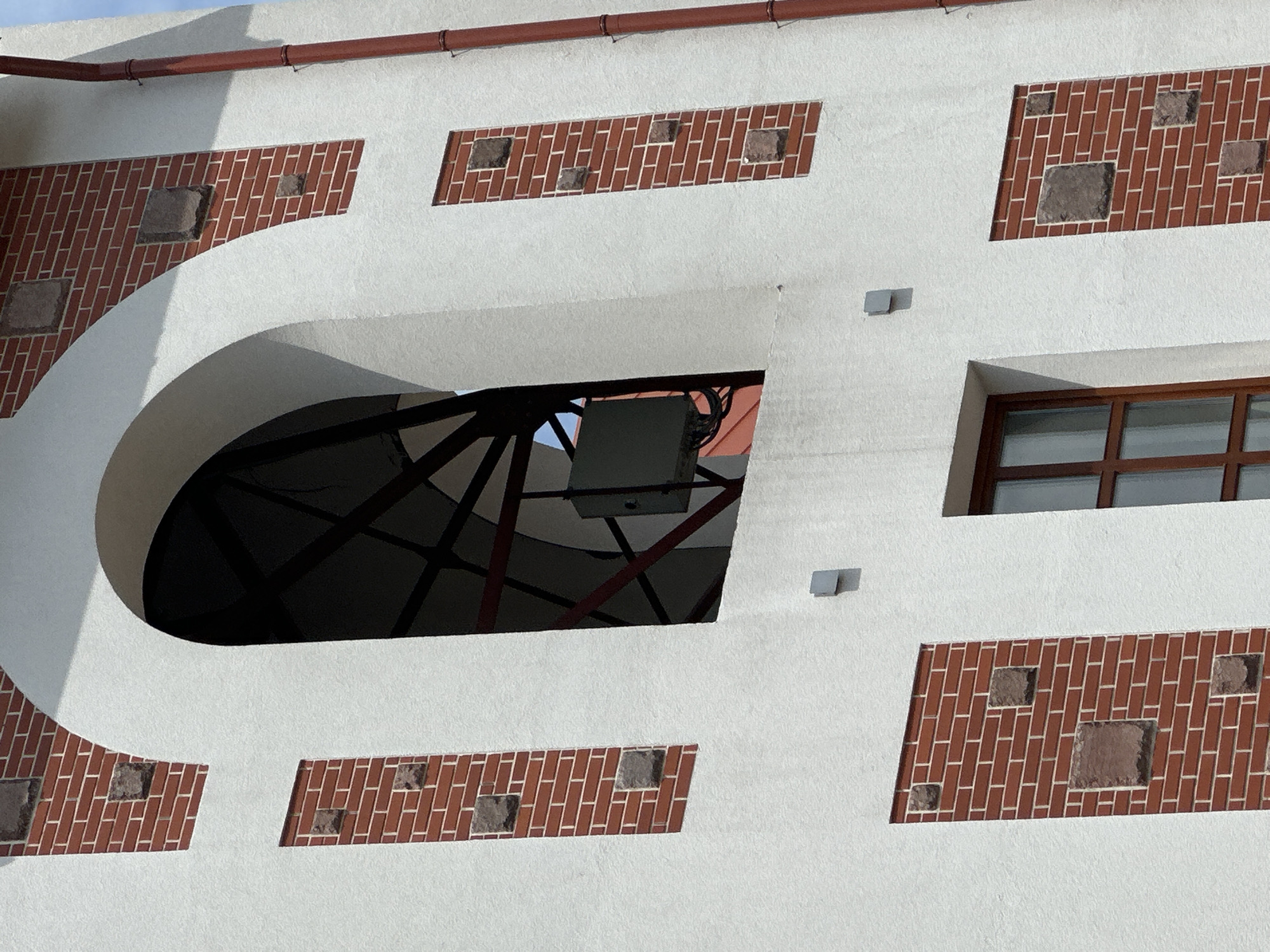 The image displays a detailed section of a building facade, characterized by white textured plaster or stucco surfaces contrasted with decorative red brick rectangular panels. These brick panels are intermittently adorned with roughly square, darker stone inserts.
Prominently featured in the upper central portion is a large, deeply recessed arched opening, which is dark within and reveals a structural metal framework. Mounted inside this framework is a light-gray rectangular equipment enclosure, from which dark cabling is visibly trailing. Through the arch, a sliver of light-blue sky and a faint reddish-orange surface are discernible in the background.
Below the arched opening, a smaller rectangular window with a brown, multi-pane wooden frame is present. To the right side of the facade, a reddish-brown vertical downspout runs from top to bottom, secured by lighter-colored brackets. Several small, light-gray square fixtures are sparsely mounted on the white plaster sections of the wall. The scene is illuminated by bright daylight, casting subtle shadows.
This architectural detail is observed on a building located in Бараўлянскі сельскі Савет, Belarus.