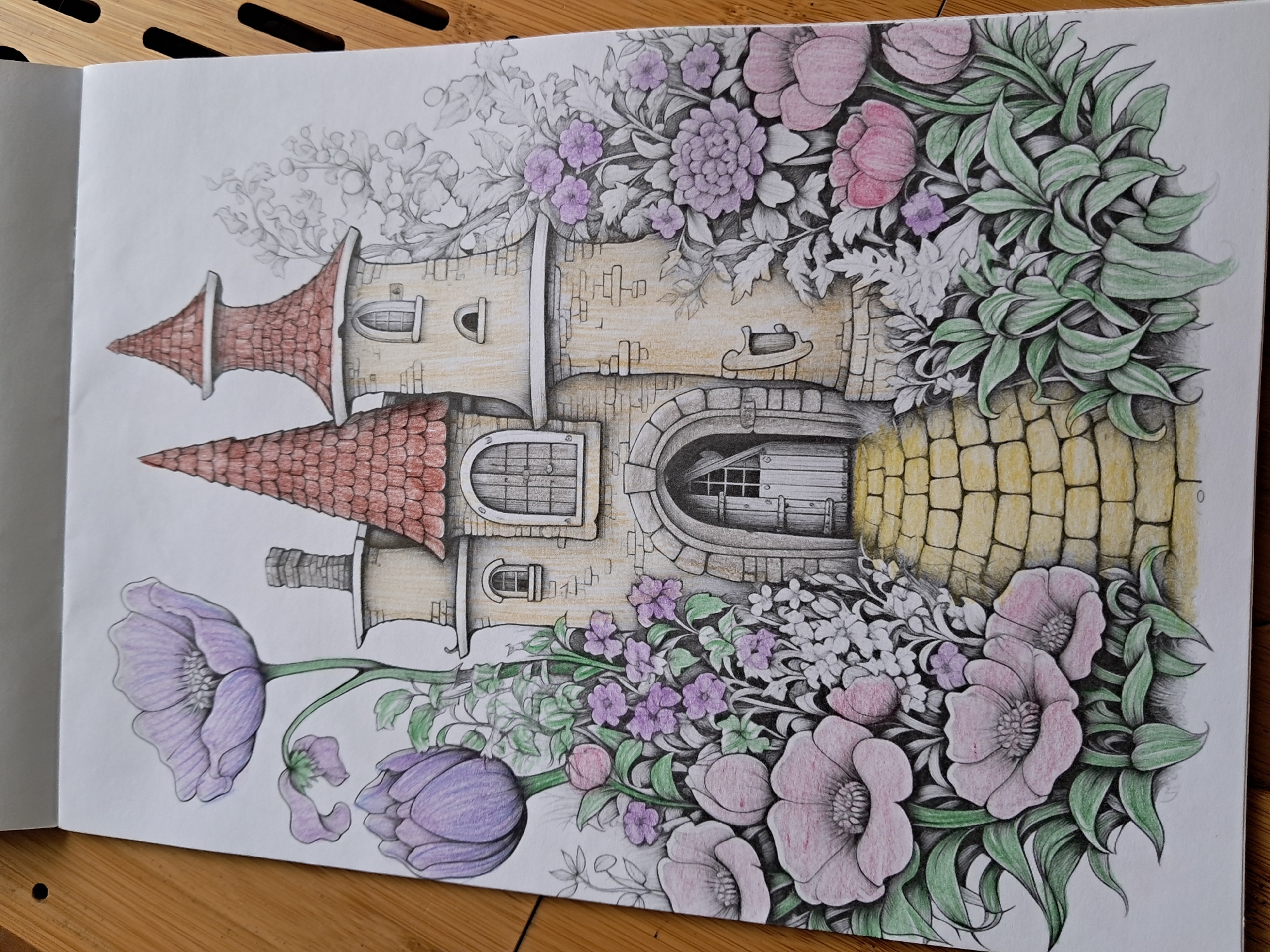 The image depicts a colored pencil drawing of a whimsical, storybook-style castle nestled amidst an abundance of colorful flowers and lush greenery. The castle has a main tower with a pointed red roof and a smaller turret with a similar roof. The stonework of the castle is detailed with brickwork and arched windows. A cobbled pathway leads to the arched entrance of the castle, which features a wooden door. Surrounding the castle are various flowers, including large pinkish-purple anemones, vibrant purple tulips, and clusters of smaller purple and pink flowers, all rendered with soft, blended colors. Green foliage and leaves fill the spaces between the flowers and the castle, creating a dense, enchanted garden setting. The drawing is presented on white paper, with the edges of the paper and the wooden surface it rests on visible in the background. The location is not specified.
