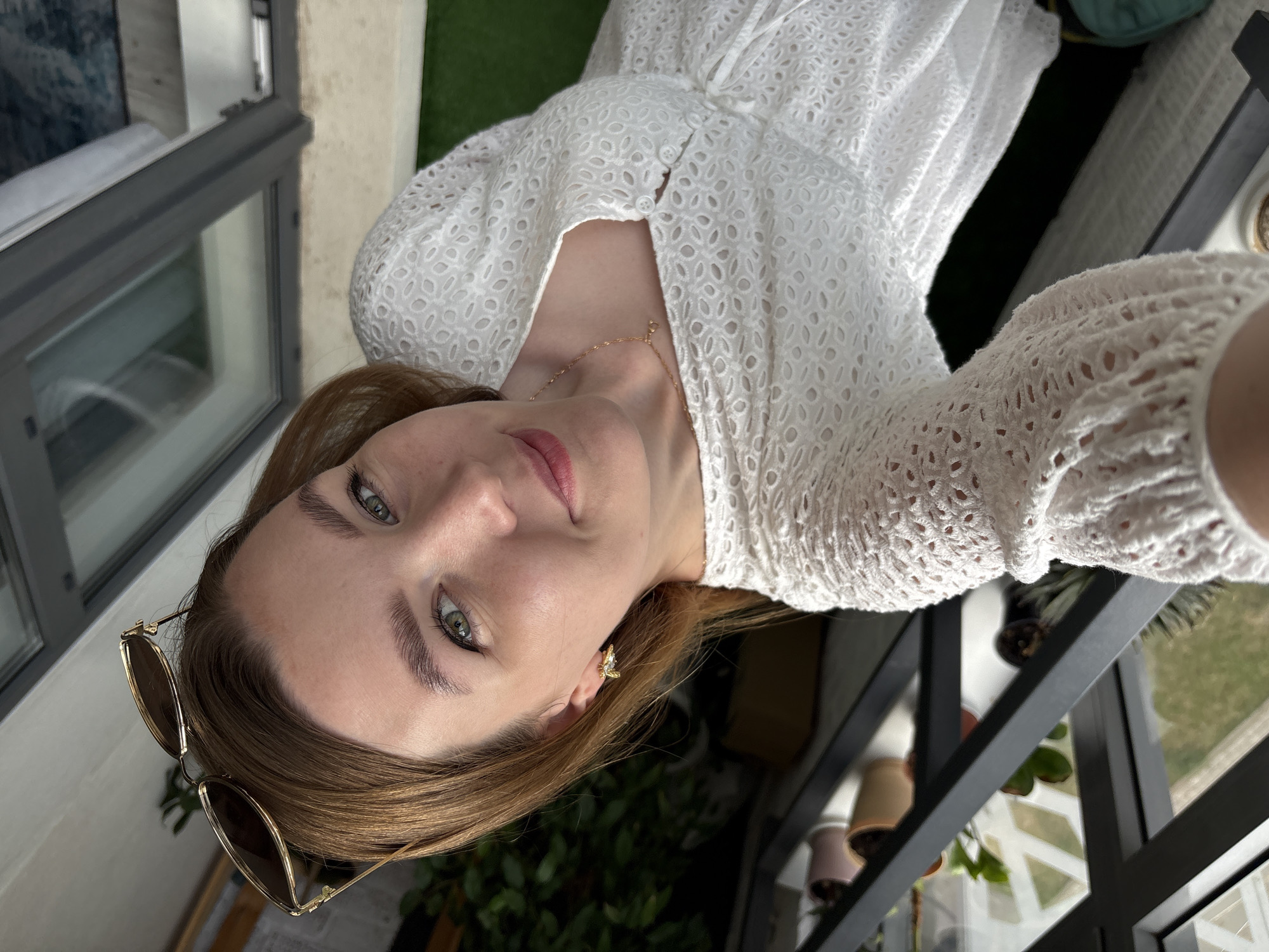 A young woman with blonde hair and green eyes is taking a selfie in what appears to be a greenhouse or sunroom. She is wearing a white eyelet dress and gold bee-shaped earrings. Sunglasses are perched on her head. Behind her, there is a rack with potted plants and a skylight. The floor is covered with a light-colored material, possibly bamboo or a rug, and a patch of artificial turf is visible. The scene suggests a bright, indoor setting, possibly a home or a commercial establishment. There is no specific information in the image to indicate the location is Izhevsk, Russia.