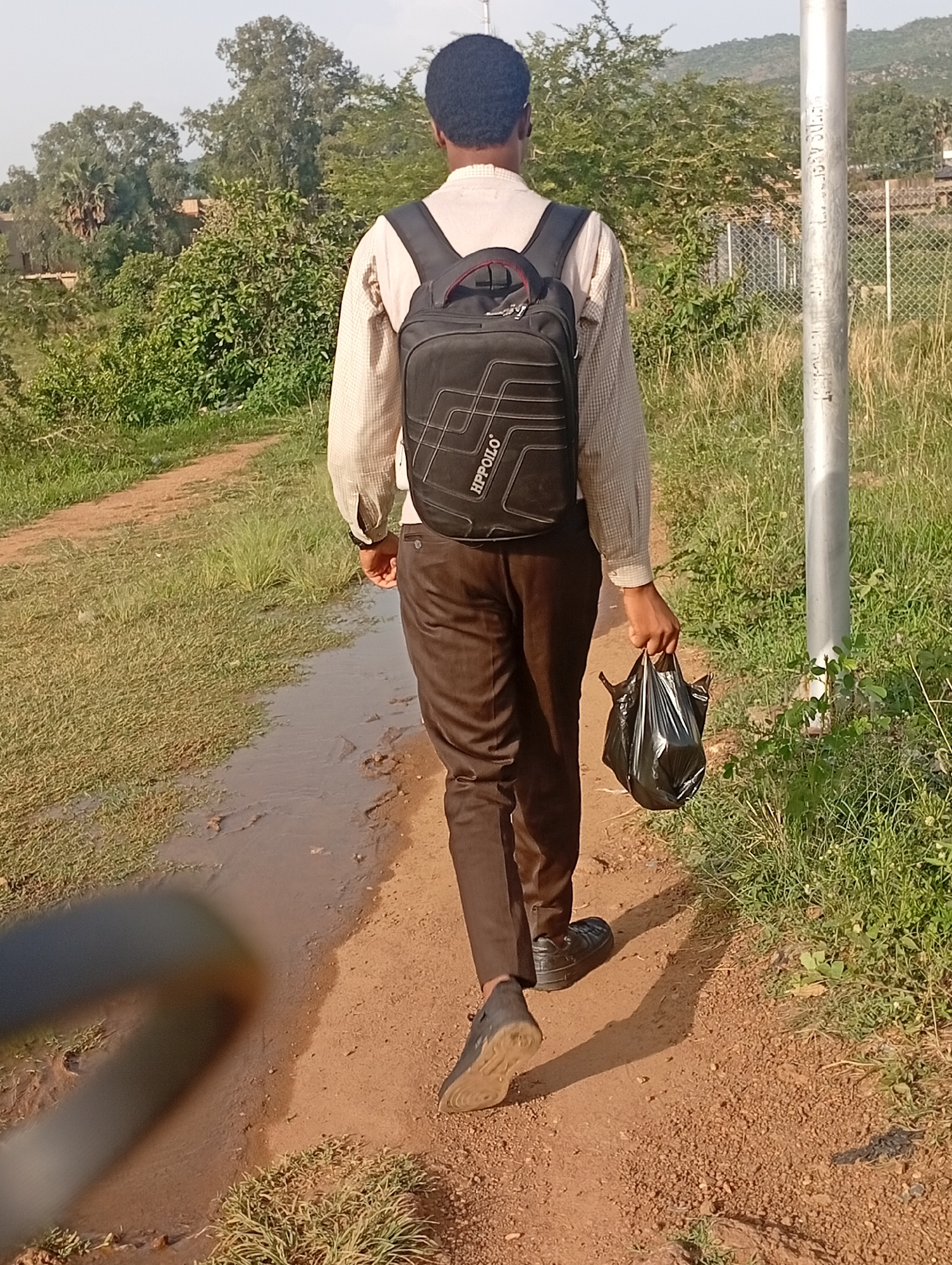 The image depicts a young man from behind, walking on a dirt path in an outdoor environment. He is wearing a light-colored, long-sleeved shirt, dark brown trousers, and dark shoes. A black backpack with white geometric patterns and the "HP POLO" logo is on his back. He is carrying a black plastic bag in his right hand. The path appears to be wet in some areas, suggesting recent rain or a puddle. The surrounding environment is characterized by dry grass, green bushes, and trees, with some buildings and hills visible in the background. The scene suggests a rural or semi-urban setting, possibly on the outskirts of the city of Jos, Nigeria, given the vegetation and terrain. The lighting indicates it is daytime, with the sun casting shadows.