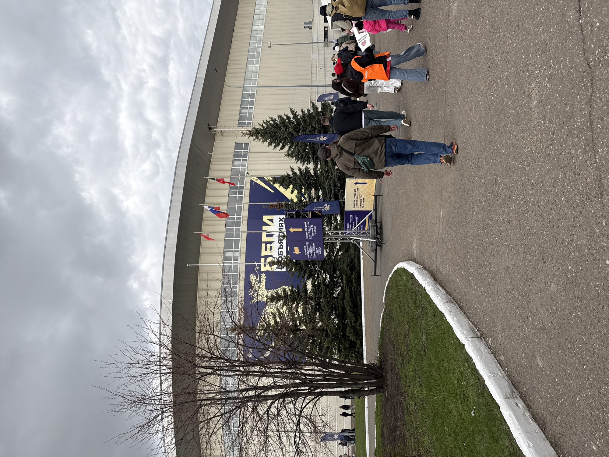 A daytime outdoor scene in Kazan, Russia, captures a queue of casually dressed people waiting in front of a large, modern building under an overcast sky. The image is oriented with the ground on the right and the sky on the left, suggesting a rotation from its natural perspective. The setting includes an asphalt paved area where people are standing, a strip of green grass with a white concrete curb, and various trees including several evergreen conifers and a bare deciduous tree in the foreground.
The background features a large, light-colored building with multiple windows and a curved roofline. Prominently displayed near the building's entrance are several large blue banners with yellow graphics and Russian text. These banners include the word "БЕГИ" (RUN) and indications of race distances like "3 KM / 10 KM / 21 KM," strongly suggesting a running event or marathon. Other textual elements on the signs and banners translate to "КАМЕРА ХРАНЕНИЯ" (LUGGAGE STORAGE) and "ПИТАНИЕ ПО ТАЛОНАМ" (MEALS BY VOUCHER).
Among the people in the queue, one individual wears a bright orange high-visibility vest with the word "ВОЛОНТЕР" (VOLUNTEER) printed on it, indicating event staff. Flags are visible on poles near the building: one Russian flag (red, white, blue horizontal stripes) and another with green, white, and red horizontal stripes (possibly the flag of Tatarstan). The people appear to be calmly waiting, likely for registration, bib pickup, or entrance to the event venue. The cloudy sky and bare tree suggest either early spring or late autumn weather conditions.