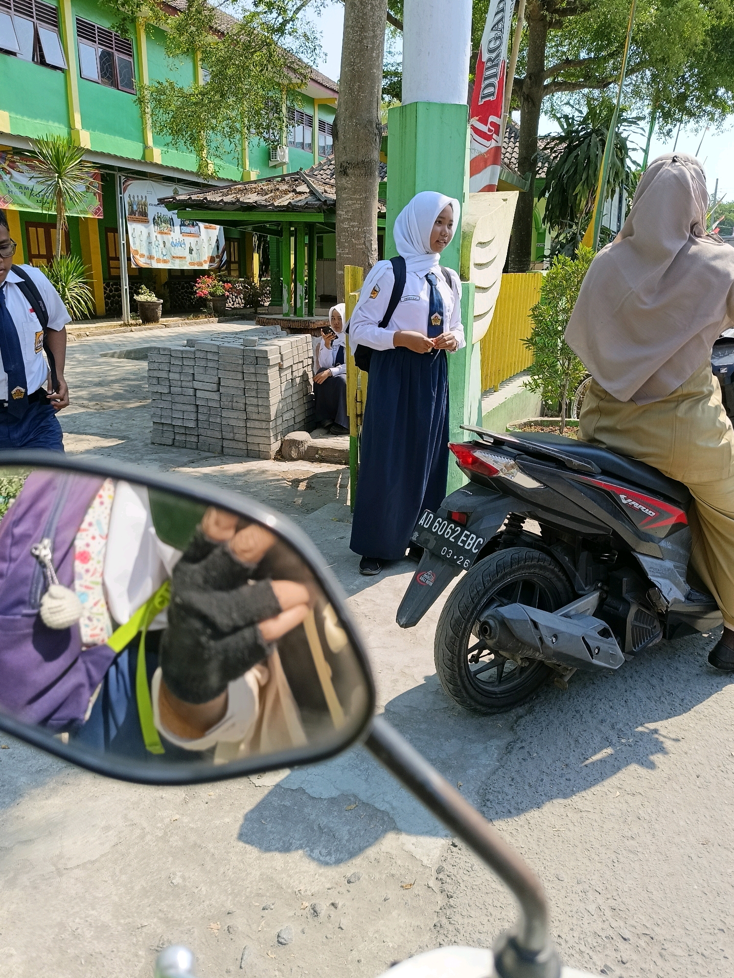 A low-angle, slightly blurred, first-person perspective shot captures a street scene outside a school building. In the foreground, a motorcycle mirror reflects a person wearing black gloves and a backpack. The motorcycle itself, black with red accents, is parked on a paved surface and has a license plate reading "AD 6062 EBC 03-26". In the background, a two-story building with a green facade and yellow trim, likely a school, is visible. Several students in white shirts and blue skirts or pants are present. One student, wearing a white hijab, stands near a yellow fence, holding a phone. Another student is seated on the ground near a stack of grey bricks. The environment appears to be a sunny day in Barukan, Indonesia, with trees and foliage adding to the outdoor setting.