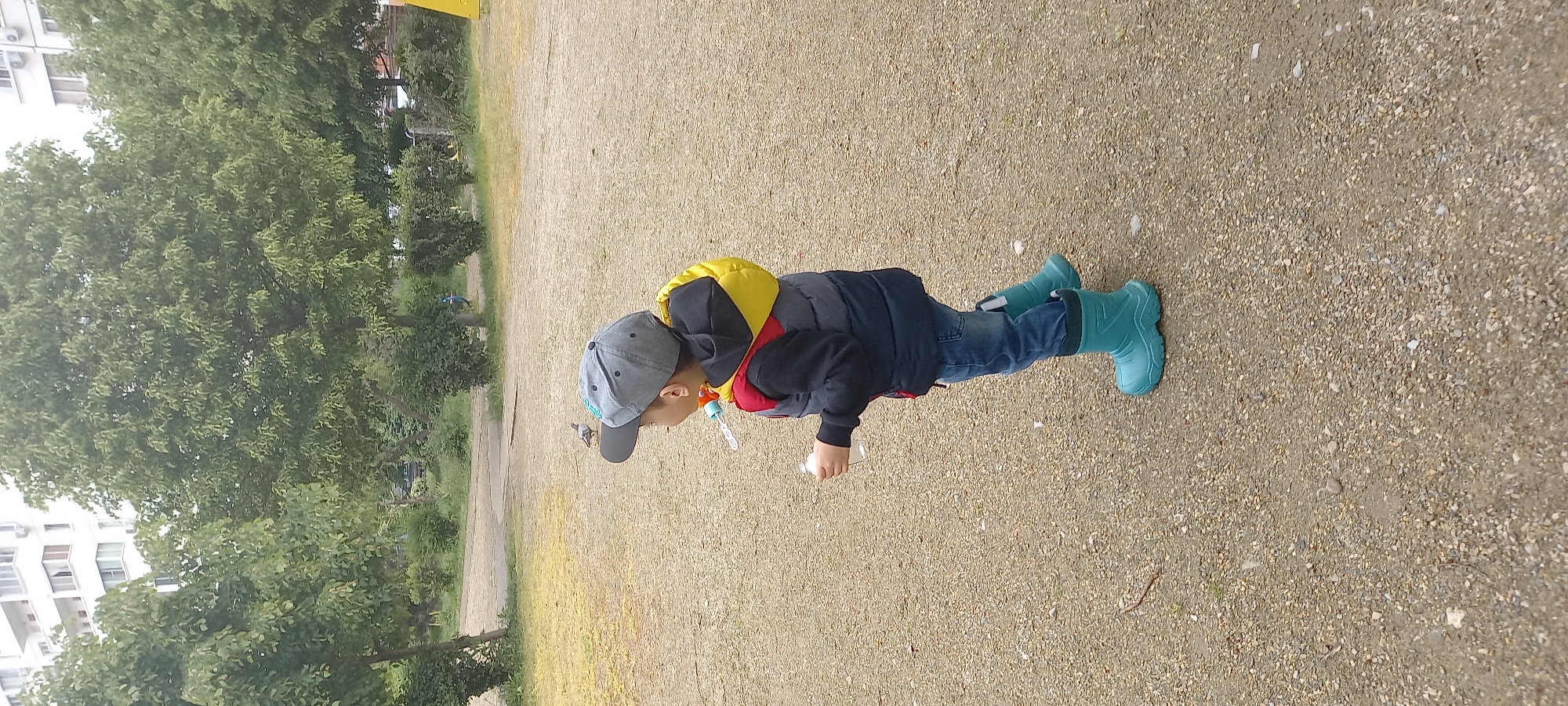 A child in a grey baseball cap, navy jacket with yellow and red hood, blue jeans, and green rubber boots stands on a gravel surface. The child holds a white container. Trees are to the left, and a building is visible in the background. The location is likely in Krasnodar, Russia.