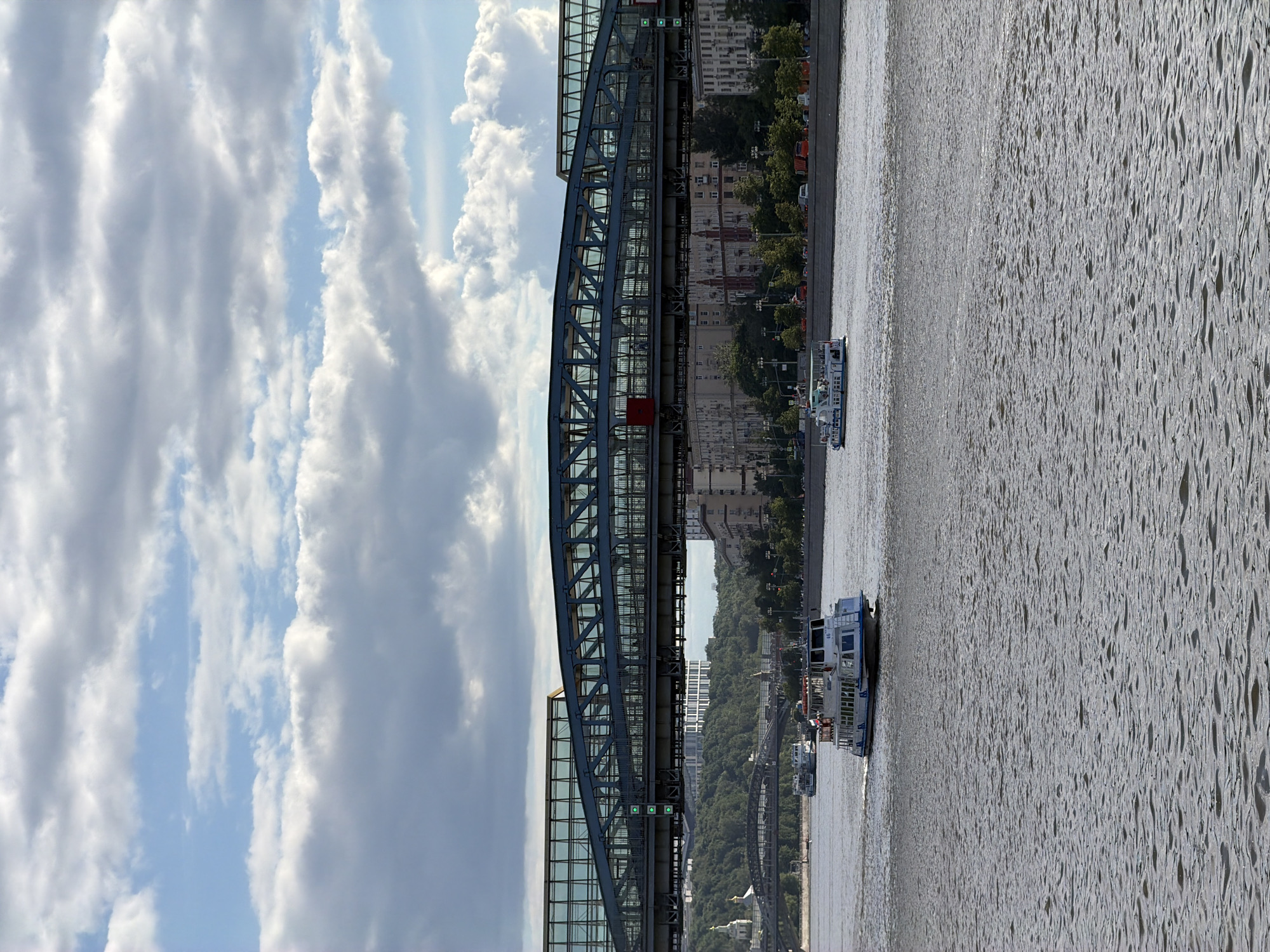 The image shows the Andrei Bridge in Moscow, Russia, spanning the Moscow River. The river flows from the bottom to the top of the frame, with visible ripples on the water surface. Two passenger boats are traveling along the river. The bridge's steel structure is prominent, with a walkway enclosed by glass panels. Behind the bridge, buildings and greenery line the riverbank. The sky is filled with white and gray clouds.