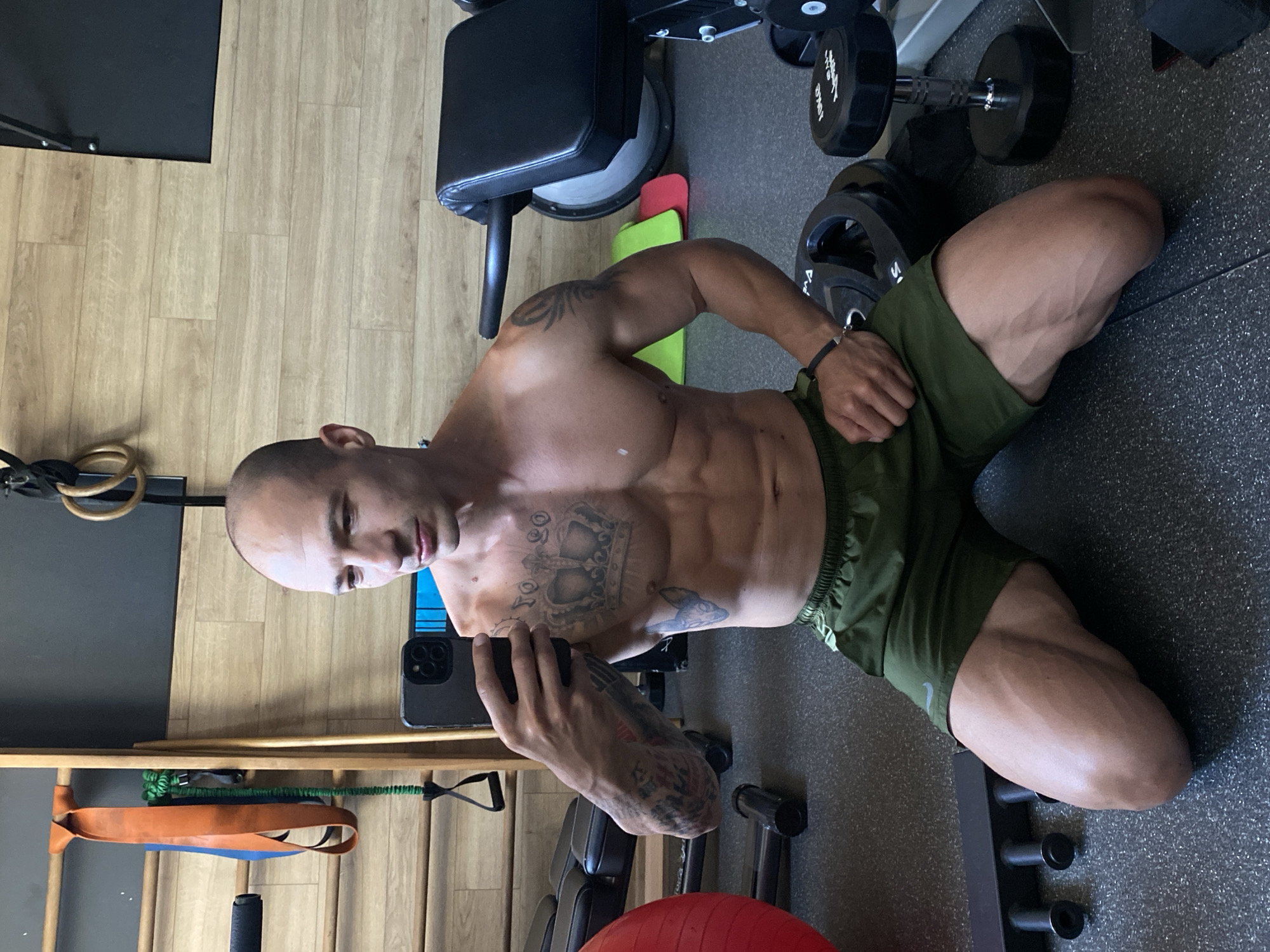 The image shows a man in a gym, taking a mirror selfie. He is shirtless and wearing green athletic shorts. He has visible abdominal muscles and tattoos on his chest and arms. He is kneeling on the floor, holding a smartphone to take the picture. In the background, there are various gym equipment, including weights, a workout bench, and resistance bands hanging on a wooden structure. The walls are paneled with light-colored wood. The gym appears to be indoors, with a dark gray floor. The man's physique suggests he is engaged in regular physical training. The image does not contain any specific elements that would identify its location in Pleven, Bulgaria, or any other specific city.