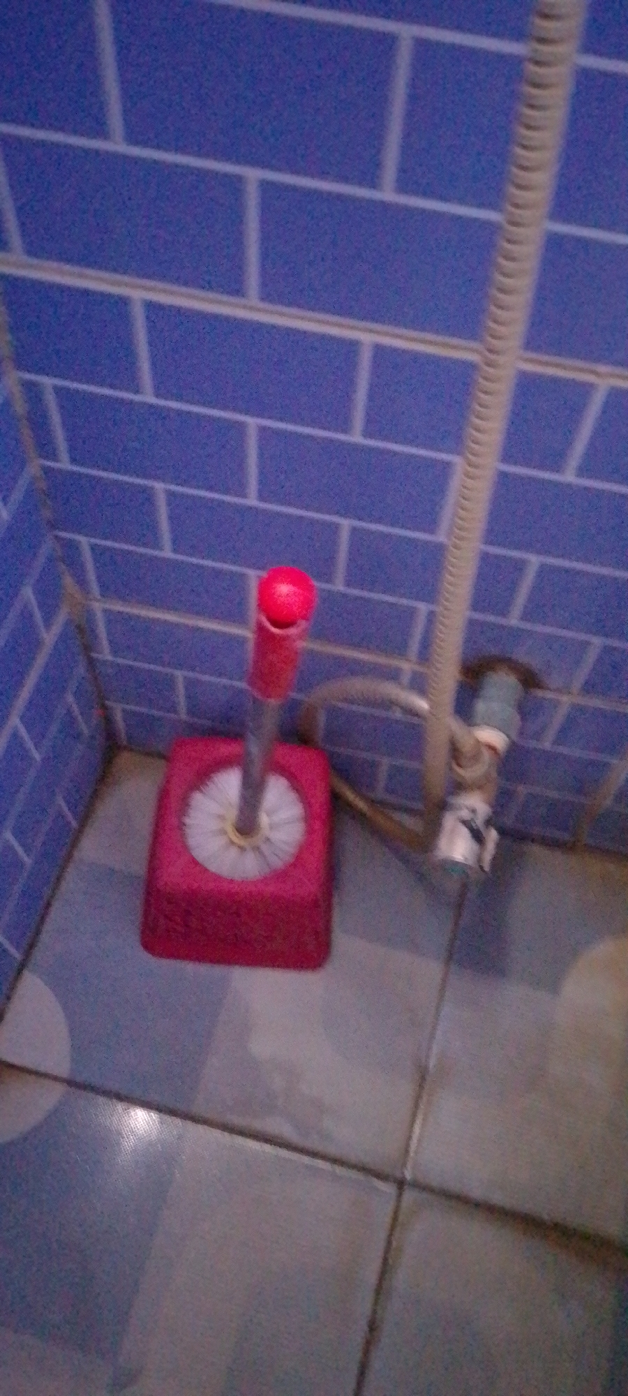 A red toilet brush with a white brush head rests in a red square holder on a tiled floor. The holder is positioned in the corner of a bathroom with blue tiled walls. A ribbed silver hose extends from the wall above the holder, connecting to plumbing fixtures. The floor tiles are a mix of blue and grey squares. The image appears to be taken at eye level and is slightly blurry.