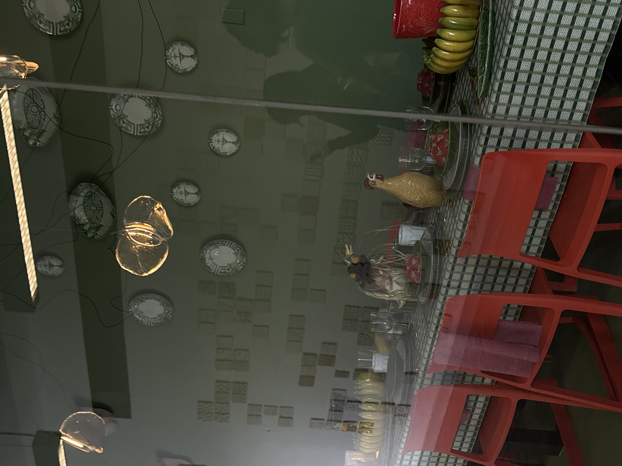 The image presents a layered scene captured through a highly reflective surface, likely a window or glass partition, looking into an interior space. This reflection overlays elements from the viewer's side onto the indoor environment.
Within the interior, a dining or display setup is visible, dominated by red accents. Several red plastic chairs are arranged around a table, which is covered with a green and white checkered tablecloth. The table is set with plates and various glassware, including stemmed and tumbler-style glasses. Decorative elements on the table include a yellow ceramic chicken figurine, a bright red ceramic pot or vase, a cluster of yellow ceramic bananas, and a green leaf-shaped dish. A centerpiece made of dried foliage and small, dark berries or fruits adds to the decor. Small, folded pink or reddish fabric pieces, possibly napkins, are placed on some of the chairs.
Reflected on the glass are a dark silhouette of a person, likely the photographer, and numerous decorative plates of different sizes hanging on what appears to be a wall. These plates display intricate patterns in shades of green, white, and grey. Dark cables are also visible in the reflection, seemingly associated with the wall decorations. Two or three glass light fixtures, emitting a warm, artificial glow, are prominently reflected. The actual background wall of the interior space is a dark, muted green, textured with a pattern of subtle square or rectangular tiles in various earthy hues.
The scene is static, depicting a prepared arrangement rather than an ongoing activity. The warm light from the reflected fixtures suggests an indoor setting, possibly in the evening. No visible text is present in the image.