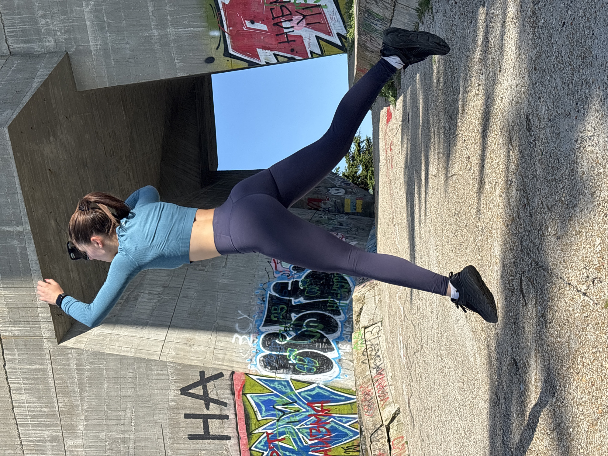 A woman in athletic wear performs a plank between two concrete structures covered in graffiti on the Asparuhovo Bridge in Varna, Bulgaria. The woman has her arms outstretched, pressed against one concrete surface, with legs extended to another. She wears a light blue long-sleeve top, dark blue leggings, and black athletic shoes. Graffiti adorns the concrete structures, including the phrase "Ha" and various abstract designs. A patch of blue sky is visible through a gap in the structure. Shadows are cast across the ground.