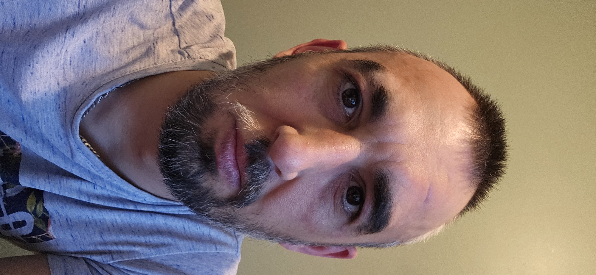 A close-up, rotated portrait shows a man with a full beard and short hair, looking directly at the camera. The image is oriented with the man's head tilted significantly, making it appear as if he is lying on his side or the photo was taken from a low, upward angle and then rotated clockwise. He has dark eyes and thick, dark eyebrows. His beard is dark with a noticeable amount of grey and white hairs, particularly around his chin and mustache. His hair, dark and short, shows signs of a receding hairline, and some grey is visible around the temples. His lips are full and have a reddish-purple hue. The man's skin tone is fair, with some minor blemishes or scars visible on his forehead/eyebrow area.
He is wearing a light blue, heathered t-shirt. A dark, patterned design, possibly floral or abstract, is visible on the lower left side of his shirt. A thin, metallic chain can be seen peeking out from under the collar of his shirt on the left side of his neck. The background is a plain, light greenish-yellow wall, suggesting an indoor setting. The lighting appears to be artificial, casting soft highlights and shadows on his face. The man's expression is neutral to slightly serious. No text is visible in the image. The photo was taken in Norwich, United Kingdom.
