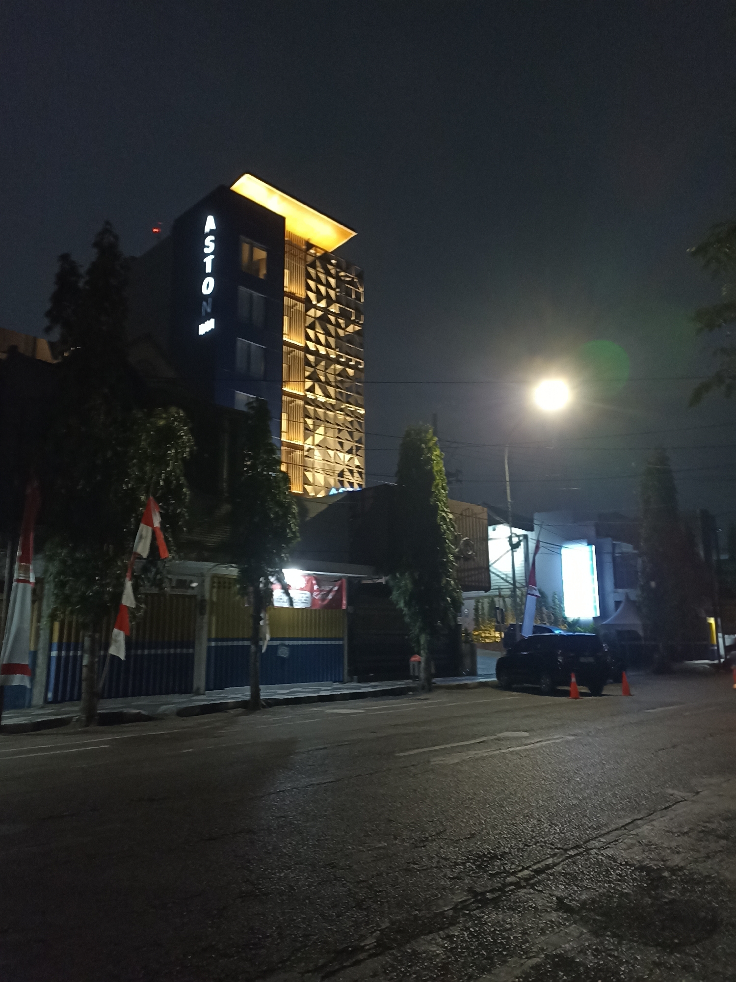 The image depicts an outdoor scene at night, likely in Lumajang, Indonesia, centered on the "ASTON INN" hotel. The hotel building, tall and modern, features illuminated signage and a distinctive geometric facade with warm yellow lighting. In front of the hotel, trees with green foliage are visible. The street in the foreground is paved, with lane markings illuminated by streetlights. A dark-colored car is parked on the side of the road, accompanied by two orange traffic cones. Indonesian flags are displayed vertically on poles along the street. Buildings with glowing signs are also present in the background, contributing to the urban night atmosphere. The sky is dark, indicating nighttime.