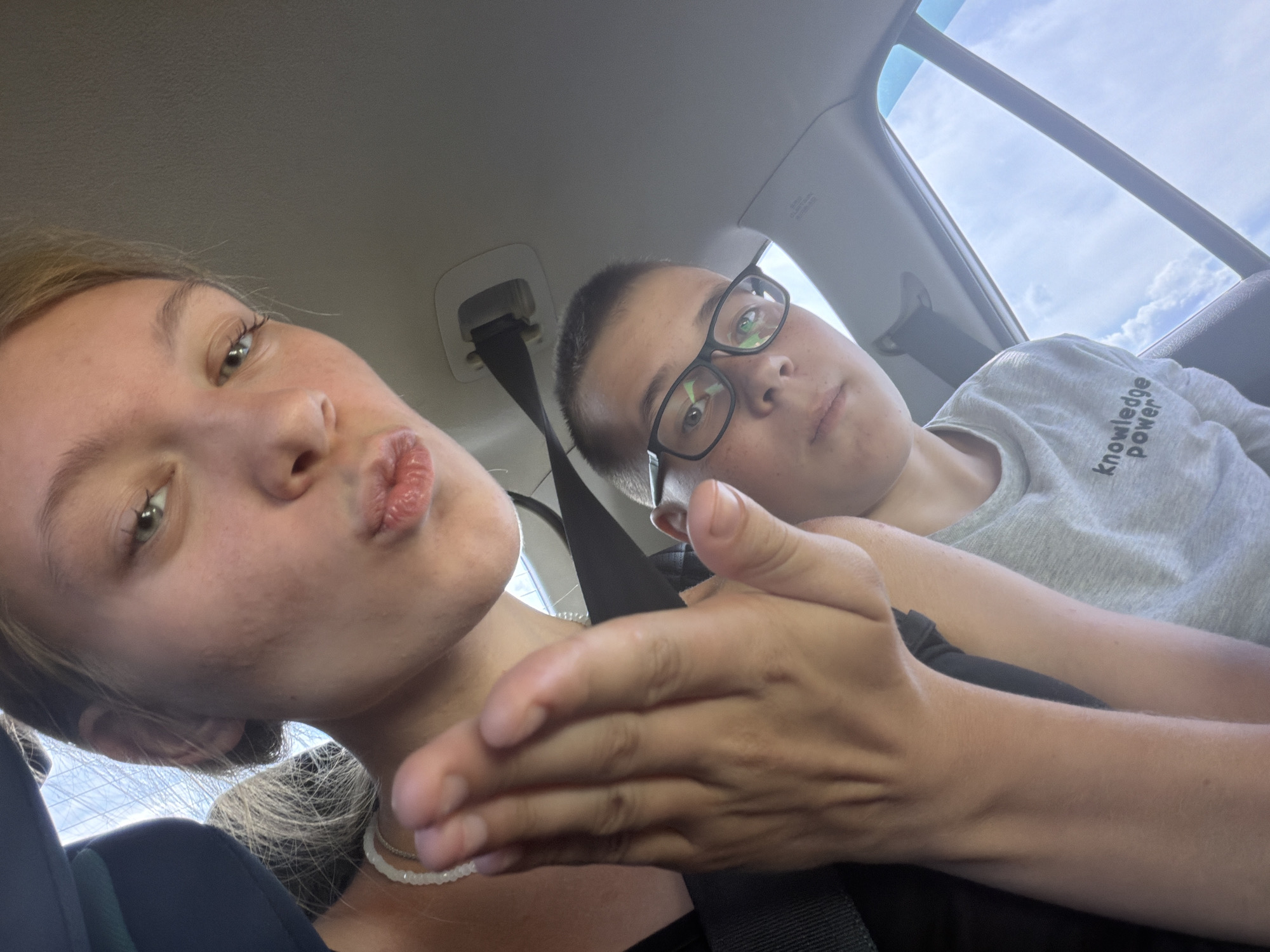 The image shows two people in a car. The person in the foreground on the left is a young woman with blonde hair, looking at the camera with her lips pursed. She is wearing a dark blue shirt and a white beaded necklace. Her right hand is extended towards the camera. To the right and slightly behind her is a young person wearing glasses and a grey t-shirt with the words "knowledge power" printed on it. They are looking away from the camera. The background shows the interior of a car, including seatbelts and the sky with clouds visible through the window. The image appears to be a selfie taken in the car, with no discernible connection to Novoe Feizullovo, Russia.
