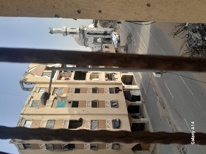 The image depicts a damaged urban environment in Khan Yunis, Palestinian Territory, viewed through two blurry, dark vertical bars in the foreground, with a light-colored wall displaying impact marks (circular holes) visible on the right.
In the midground, a multi-story building, light beige with brown trim, stands significantly damaged. Its upper floors exhibit extensive structural damage, including missing sections and exposed interiors. Numerous windows are broken or missing, some covered with blue tarps or dark materials, and clothes hang from a lower-level window. The building's ground floor features arched openings.
A paved road occupies the central midground, with a double solid white line and scattered debris. A gray streetlamp pole stands centrally, partially obstructing the view.
To the right, beyond the streetlamp, a white mosque with a dome and a partially damaged minaret is visible. Adjacent to the mosque, on the street, a small white tent structure is erected, surrounded by approximately four individuals. A white vehicle is parked nearby. Another lone individual is visible walking further down the street to the right.
The bottom right foreground contains a pile of concrete rubble and twisted rebar. The sky above is clear. The bottom left bears a "Galaxy A14" watermark.
