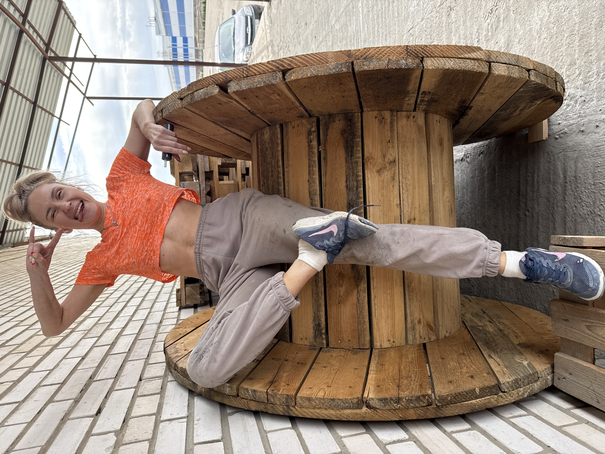 The image depicts a woman lying under a large wooden cable spool, located outdoors. The woman, with blonde hair tied back, wears an orange cropped t-shirt, grey sweatpants, white socks, and blue sneakers. She is smiling and making a "peace" sign with her left hand. The cable spool is constructed from wooden planks and sits on a brick-paved surface. In the background, there's a view of a building with blue and white striped roofing and a silver car. The scene is in Chaikovsky, Russia.