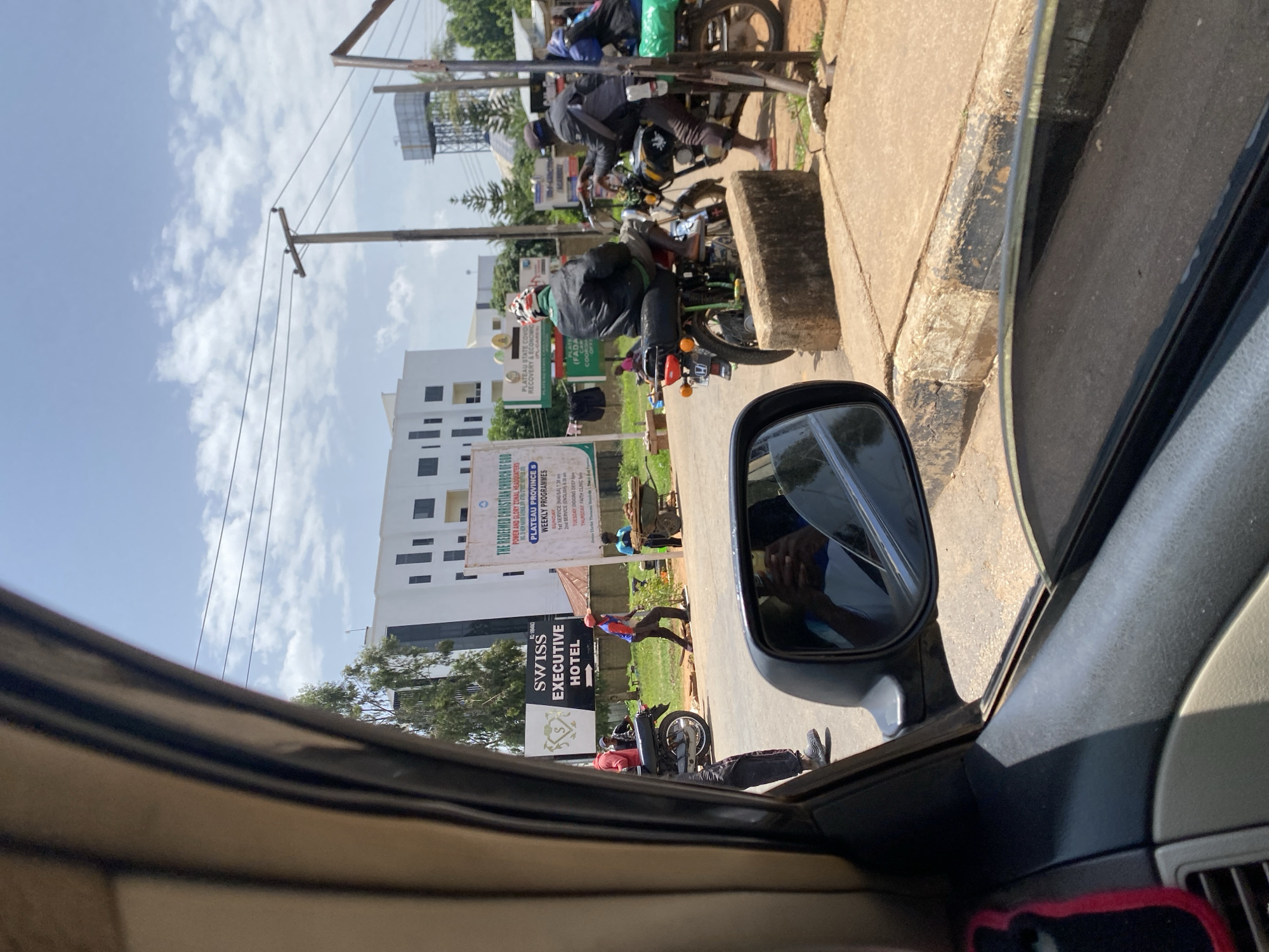 The image captures a street scene in Bar Jirgi Summa, Nigeria, viewed from inside a vehicle. In the foreground, the interior of the car, including a side mirror reflecting a person holding a phone, is visible. Outside, a paved road and sidewalk are present. Several people are seen on or near motorcycles. One motorcycle, a red and black model, is parked on the left. Further down the road, two men are on a black motorcycle, with one appearing to be adjusting something on it. Another motorcycle, with a green bag strapped to the back, is positioned to the right. Behind the people and motorcycles, a large white building with multiple windows stands, and a sign that reads "SWISS EXECUTIVE HOTEL" is visible. To the right of the hotel sign, a green and white banner displays text, likely advertising a church or community event. In the background, trees, utility poles, and a water tower are present under a partly cloudy sky. The overall environment suggests a moderately busy urban or suburban area.