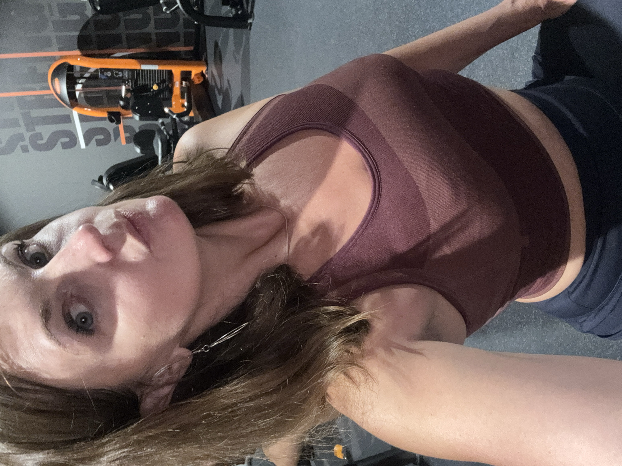 A woman is taking a selfie in a gym. She is wearing a burgundy sports bra and dark leggings. She has brown hair and is wearing dangling earrings. The background shows gym equipment, including an orange weight machine, and the floor is dark gray. There is text on the wall behind her. The image appears to be taken indoors in a well-lit environment. There is no information in the image to suggest the location is Saint-Claude, France.