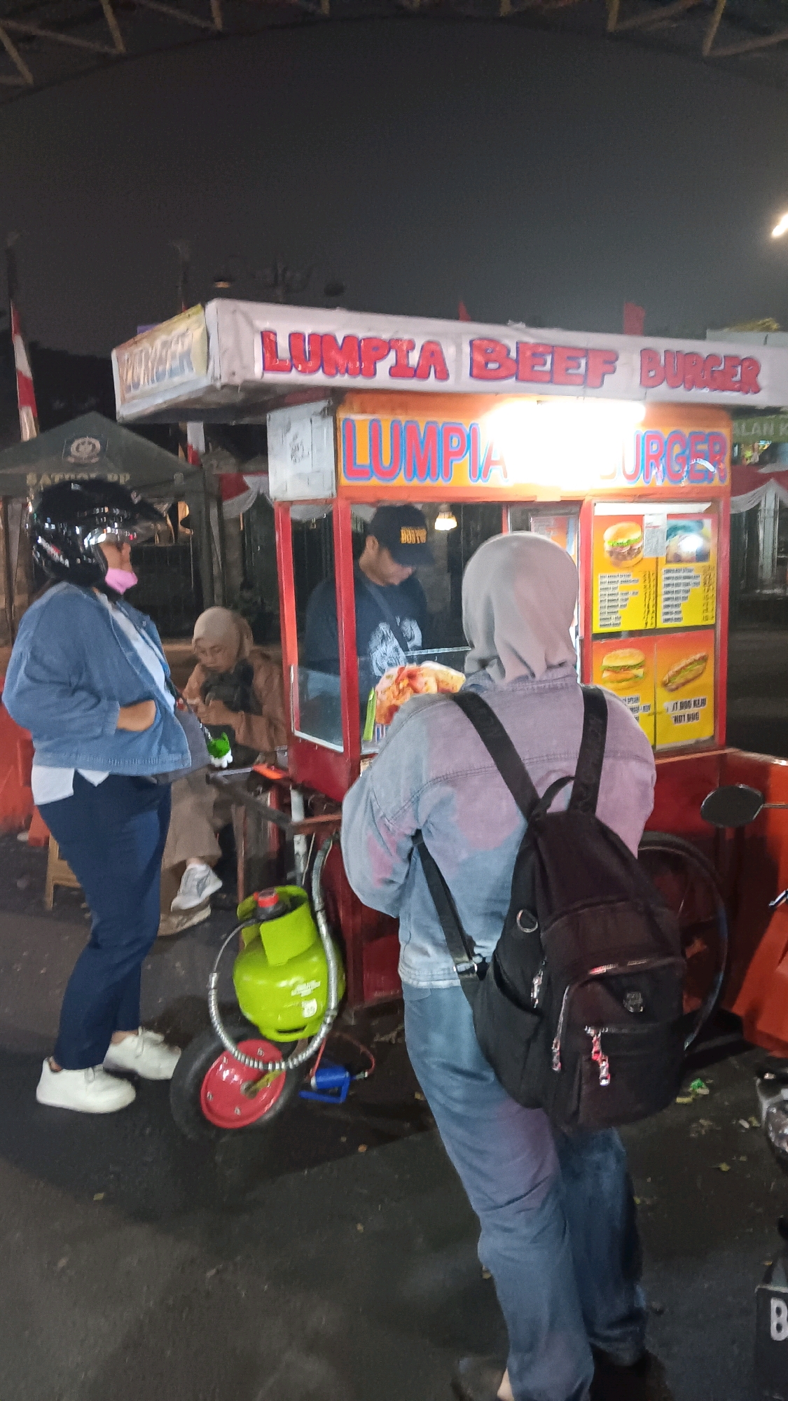 An outdoor night scene in South Jakarta, Indonesia, features a food stall that sells "Lumpia Beef Burger." The stall has a red and white striped awning and a menu board displaying pictures of burgers and hot dogs. Several people are present: a woman wearing a denim jacket and helmet stands to the left, facing the stall; another person, also wearing a light-colored head covering and denim jacket, is standing at the counter of the stall, facing away from the camera; and a man wearing a black cap and t-shirt is working behind the counter. A green gas cylinder is visible next to the stall, attached to a wheeled cart. The ground appears to be asphalt, suggesting a street or parking area.