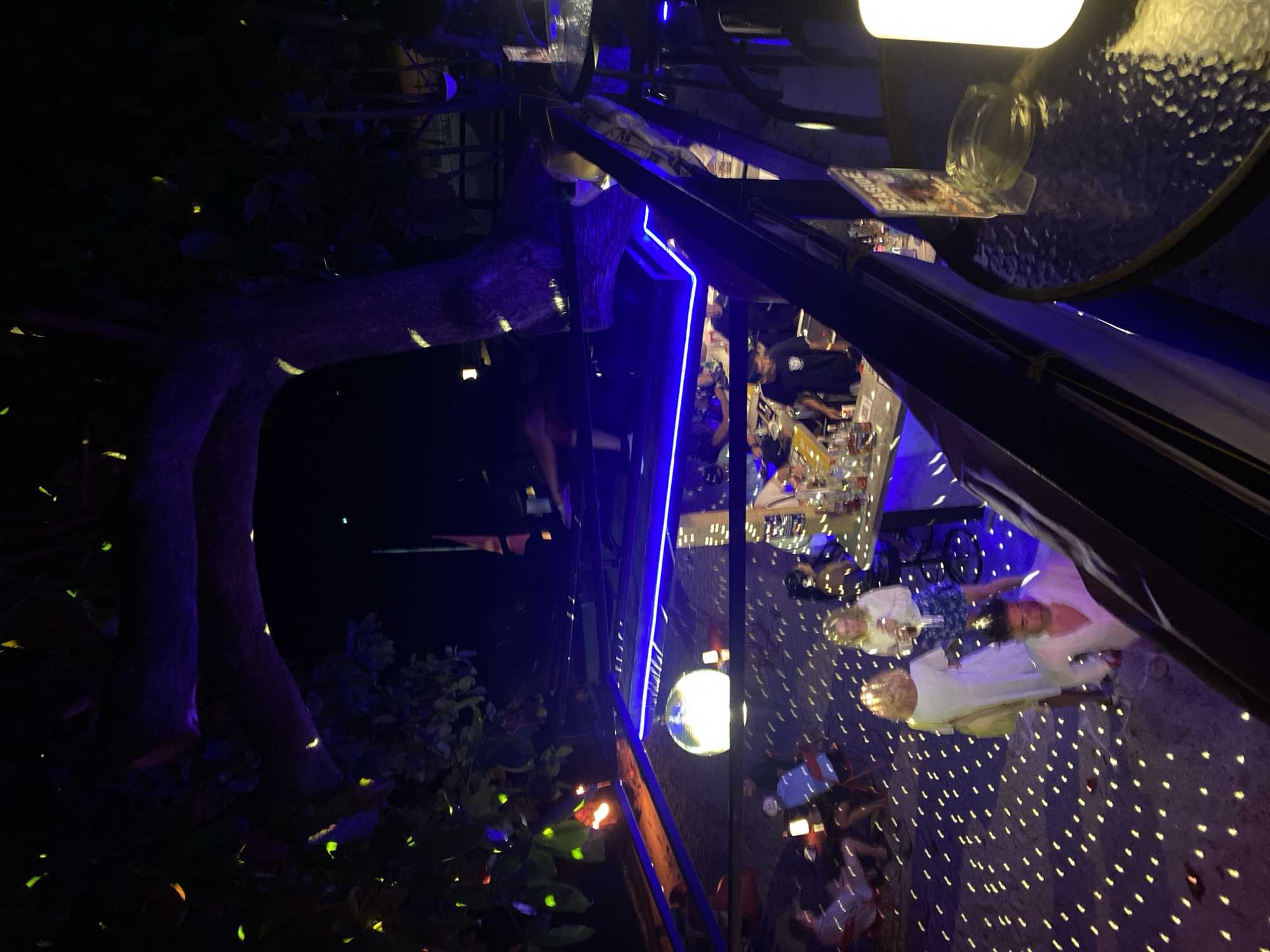 The image depicts an outdoor bar or restaurant setting at night in Ban Haad Rin, Thailand. Tables are visible, some with customers seated and interacting. There are people wearing light-colored clothing. The environment is decorated with lighting, including blue neon lights, overhead lamps, and a disco ball that projects light patterns on the ground. A tree and foliage are present outside the bar area, with some illuminated areas suggesting outdoor lighting.