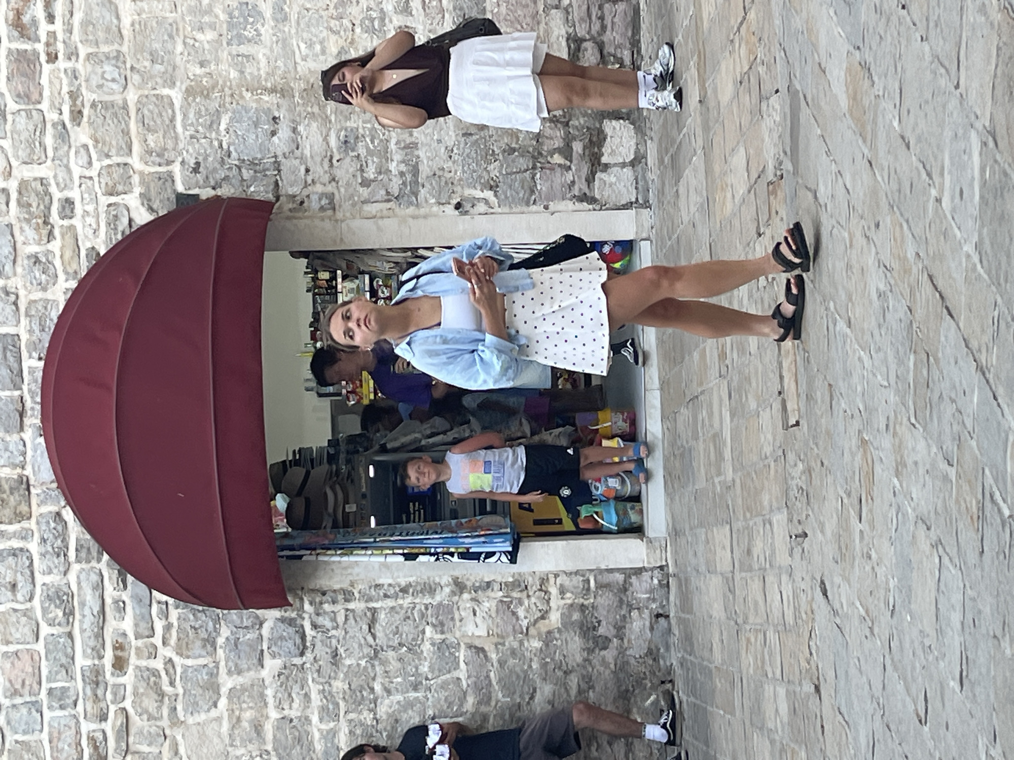 In Budva, Montenegro, a young woman in a blue shirt and a white polka dot skirt walks on a cobblestone street, looking up. She holds a smartphone in her right hand. To her right, another woman in a dark top and white skirt holds a smartphone and takes a photo. Behind them, a young boy in a gray tank top and black shorts stands in the doorway of a shop that sells hats and other items. A man in a dark t-shirt and shorts stands to the left of the boy, holding two jars. The shop is built into a stone wall, with a maroon awning over the entrance. The street is paved with light-colored flagstones.