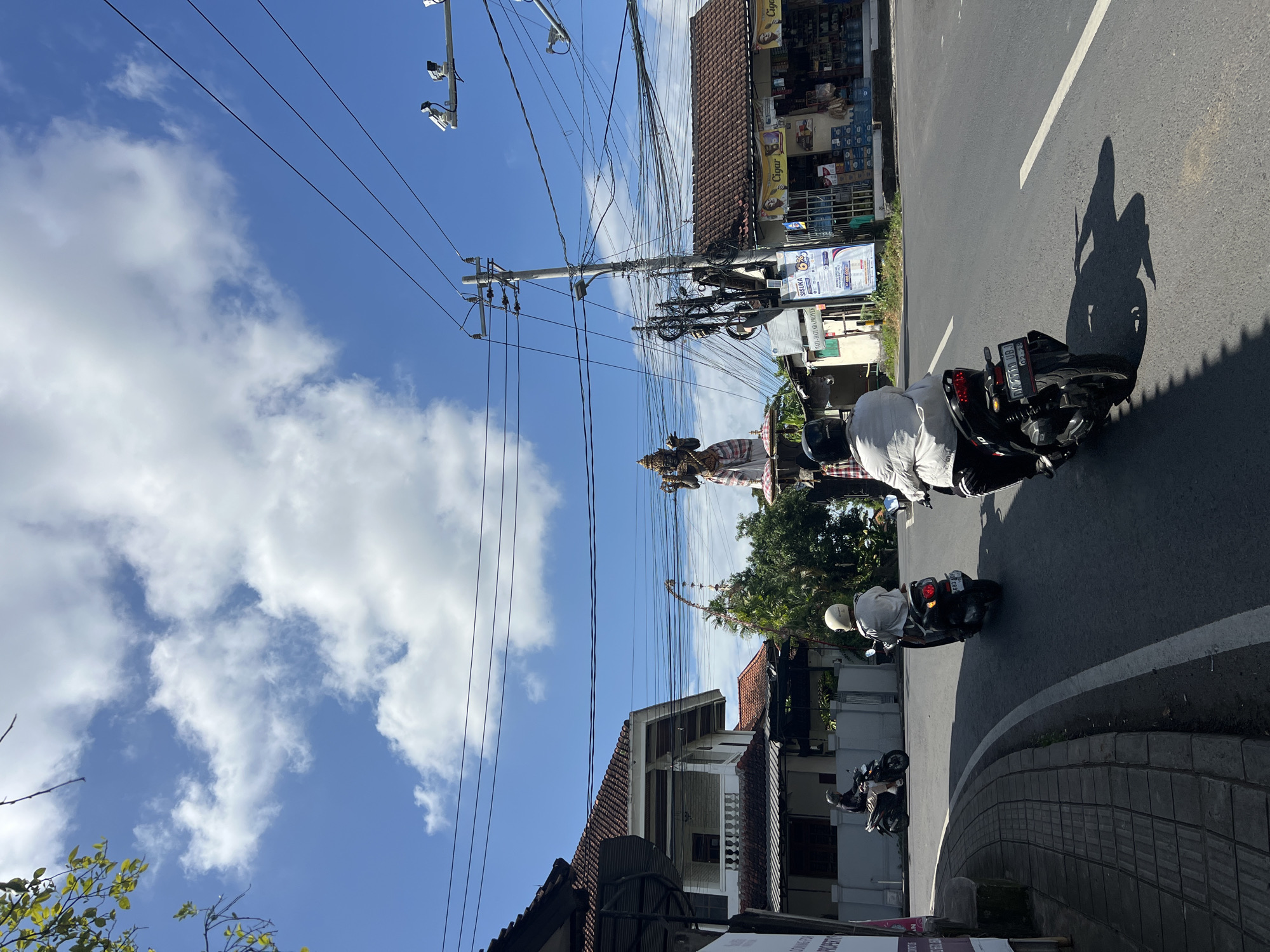 A clear daytime scene in North Kuta, Indonesia, features a road with several motorcycles. Two motorcyclists, one wearing a white shirt and the other a black shirt, are riding away from the camera on the right side of the road. A third motorcycle, with a rider in a white shirt, is further ahead on the left. A large, multi-tiered statue of a deity, possibly Hindu, stands prominently in the background, adorned with traditional Indonesian fabric. Several buildings, including a house with a white facade and a shop with a brown tiled roof, are visible along the road. Utility poles with a dense network of wires crisscross the sky, which is partly cloudy with a bright blue expanse. The road surface is asphalt, marked with white lines, and a tiled sidewalk is present on the left. The overall impression is of a bustling street scene in a tropical urban environment.
