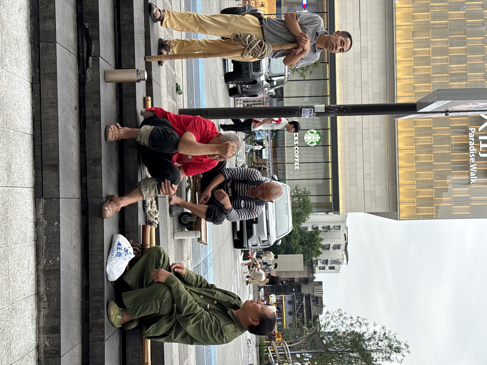 The photograph captures a candid scene on a staircase in an urban setting, likely the Nan'an District in China. Three men are seated on the stairs, with a fourth man standing above them.
One man, dressed in an olive green uniform-style outfit, sits on the lower steps with a white plastic bag next to him. A second man, clad in a red shirt and dark pants, is crouched with his head in his hands. Next to him, a third man wearing a striped shirt is seated on a small bench.
The man standing on the top stairs is dressed in a gray shirt and khaki pants. He holds a bamboo pole with rope wrapped around it. Behind these figures, the backdrop includes pedestrians, cars, and buildings. Notably, a Starbucks sign is visible, as is a sign that reads "Paradise Walk," further suggesting the location. The overall environment has the feel of a bustling city street.