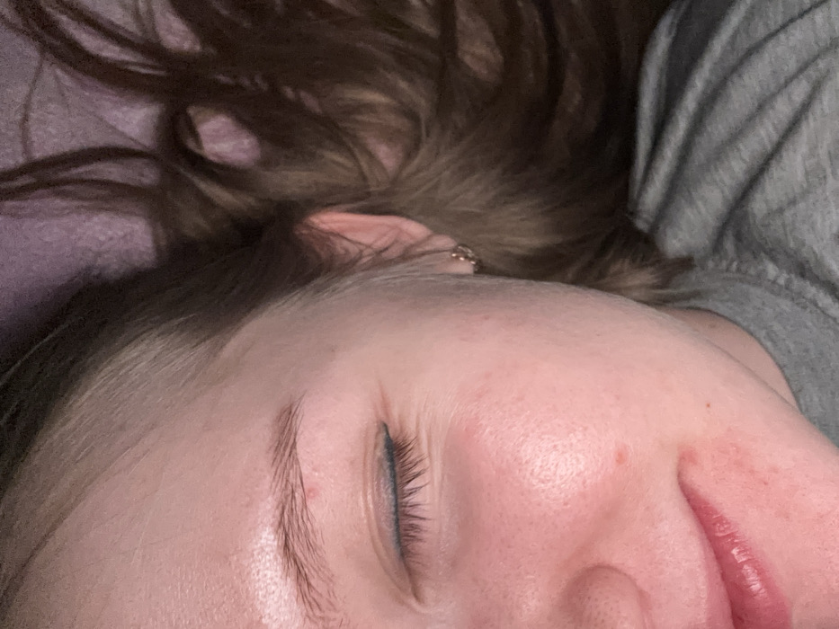 The scene depicts the right side of a female individual's face in a close-up, angled perspective, captured within an indoor environment, understood to be located in Nizhnekamsk, Russia. The subject is positioned lying down, with the right eye closed. A thin line of blue eyeliner is visible along the upper eyelid. Fine lines are present at the outer canthus of the closed eye. The right eyebrow is light brown. The skin appears light-toned with several minor red blemishes on the forehead, right cheek, and near the mouth. The lower portion of the image shows natural pink lips, slightly parted. A small, gold-colored hoop earring is present in the lobe of the right ear. Light brown hair, exhibiting lighter streaks, is fanned out against a textured, dark purple or pink surface, consistent with bedding. A grey fabric garment, identified as a t-shirt, covers the subject's right shoulder and upper chest. Illumination is soft, originating primarily from the left.