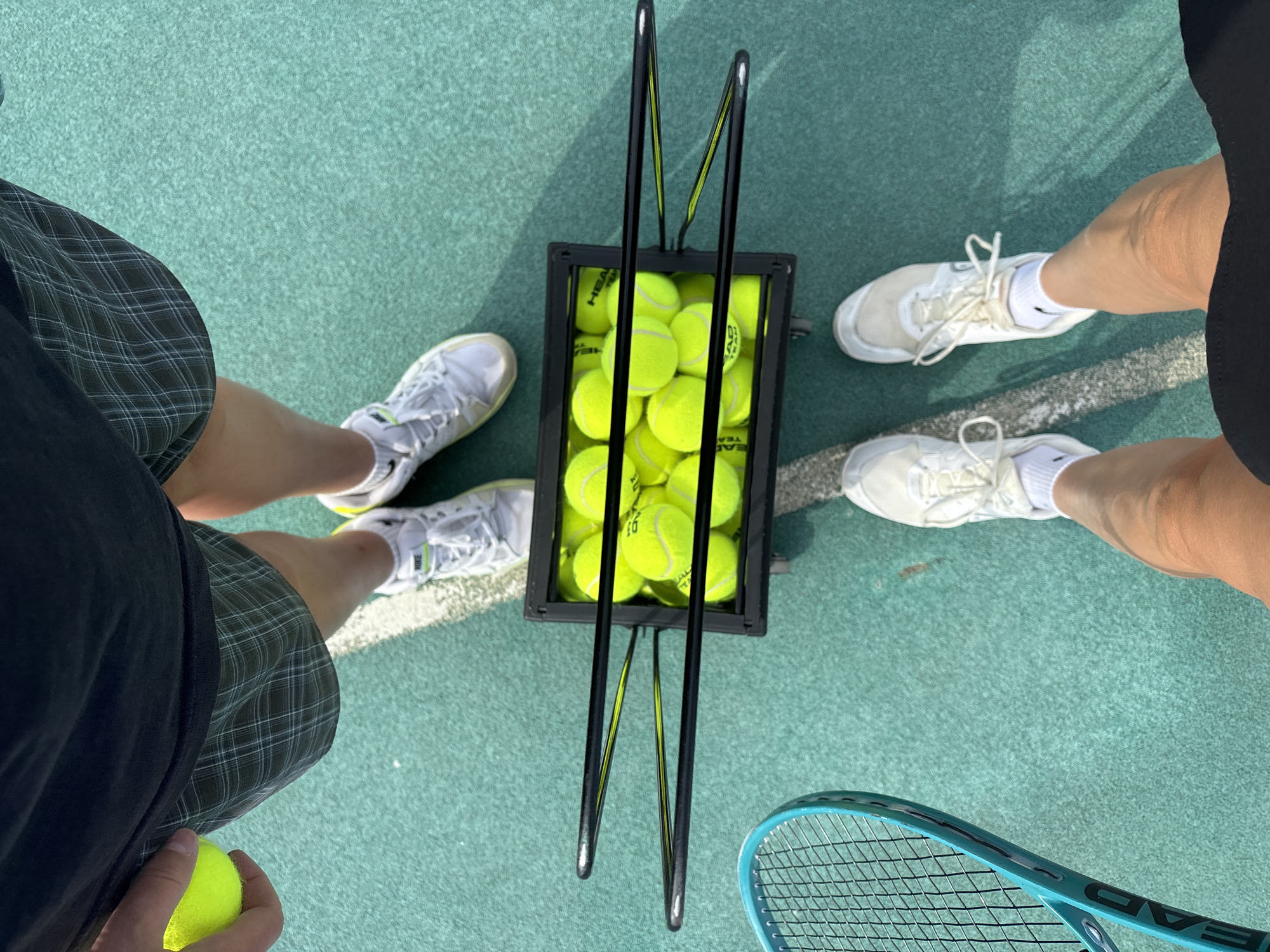 The image shows an overhead view of a tennis court. A black basket filled with yellow Head tennis balls is centrally positioned. Two individuals stand around the basket. The person on the left wears a black shirt and plaid shorts, holding a tennis ball. The person on the right is dressed in black. Both are wearing white sneakers. A blue tennis racket is visible at the bottom of the frame. The setting is outdoors on a green tennis court with white boundary lines. The location is not specified in the image and therefore can't be reliably inferred as Chaikovsky, Russia.