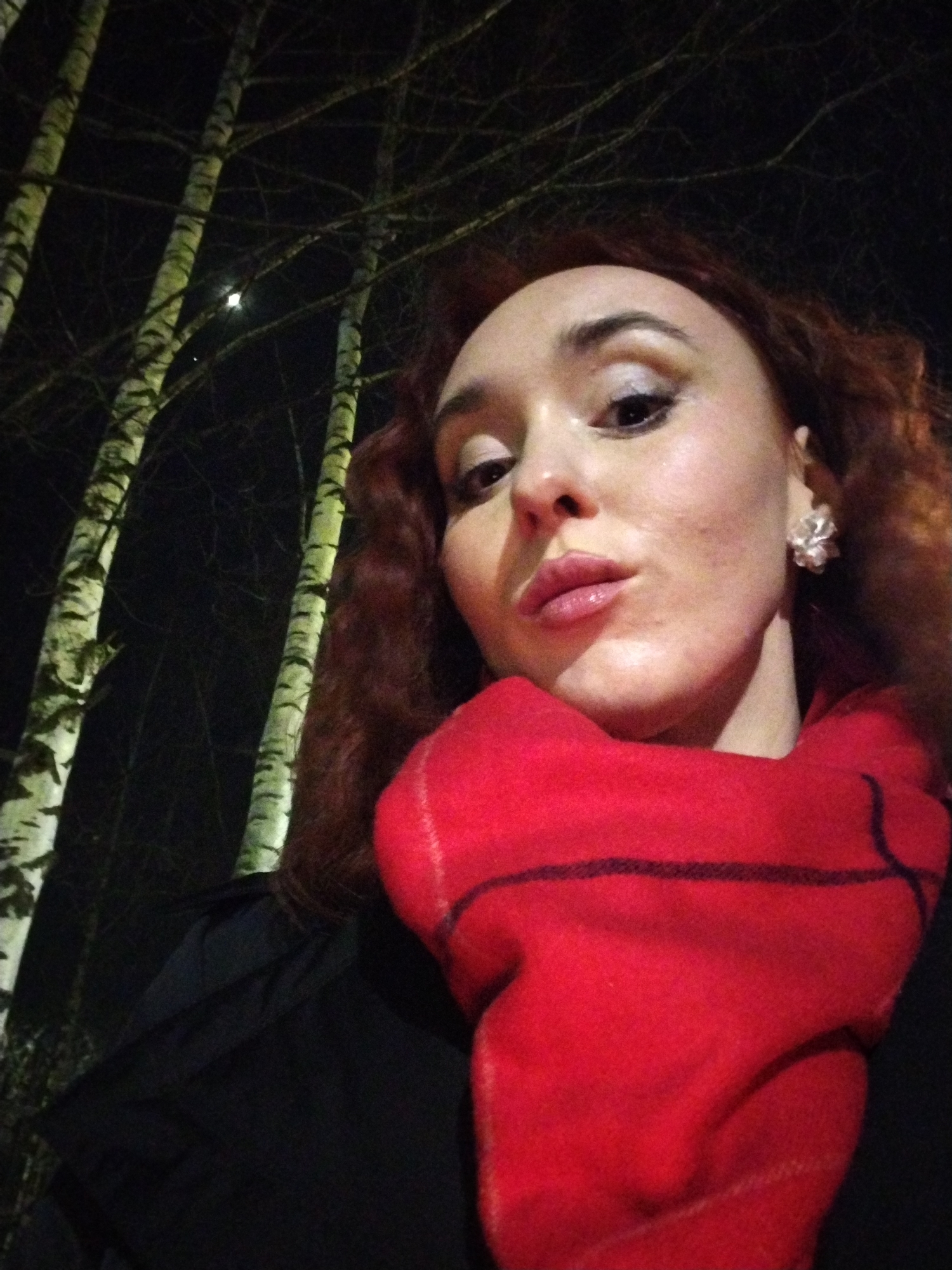 A female individual is captured in a nighttime, upward-angled selfie. She has reddish-brown, wavy hair, and fair skin with visible pores and minor blemishes. Her eyes are made up with eye shadow, and her lips are pursed, covered in light pink lipstick. A bright, multi-faceted earring is visible on her right ear. She wears a thick, red scarf with a dark, possibly plaid, pattern, wrapped around her neck, covering a dark-colored outer garment. The background is dark, indicating night. Multiple birch trees with bare branches and distinctive white and black bark are visible, illuminated from the left, suggesting artificial lighting. A small, bright light source, possibly a distant light fixture or celestial body, is visible between two tree trunks in the upper left quadrant. The scene occurs outdoors, likely in an area with natural foliage, but no specific landmarks or features are present to determine the exact location within Izhevsk, Russia.