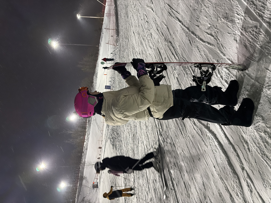 A nighttime scene depicts a snow-covered ski slope, identified as being located in Курово, Russia. The environment is illuminated by multiple tall floodlights, visible against the dark sky, under which light snow is actively falling.
In the foreground, a person stands in profile, facing right, holding a snowboard vertically with both hands. This individual wears a bright pink helmet with orange goggles resting on top, a light beige quilted puffer jacket, black cargo-style ski pants, and black snowboard boots. Dark gloves with purple and pink accents are worn on both hands. The snowboard is white with red and black graphics and features black bindings.
In the mid-ground to the left, a person wearing a yellow jacket and dark pants is visible standing on skis with poles. To their right, another individual in dark winter sports attire, including a helmet and goggles, is on skis and appears to be in motion, causing a slight blur. A smaller figure in a pink jacket is positioned between these two skiers. The background reveals additional indistinct figures on the slope, red safety netting, and snow-covered trees lining the perimeter of the ski area. The snow surface shows visible ski and snowboard tracks.