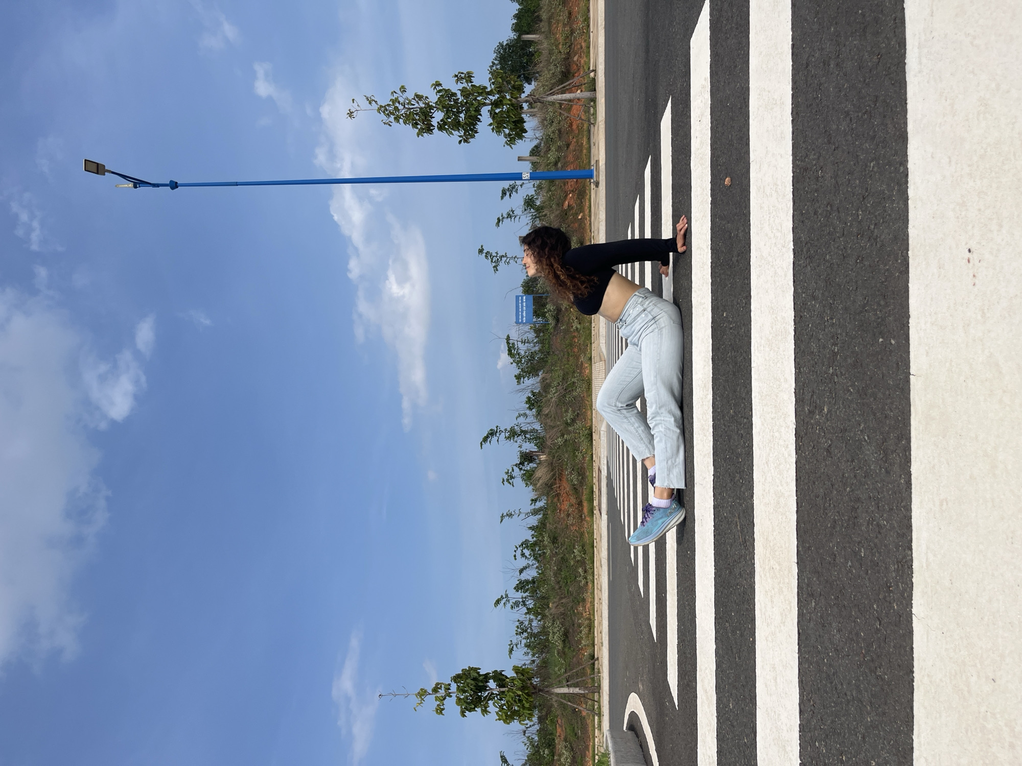 A person is lying on their back on a pedestrian crossing in Mui Ne, Vietnam. The person is wearing a black crop top, light blue jeans, and blue sneakers. Their head is tilted back and looking up at the sky. The pedestrian crossing is on an asphalt road with white stripes. In the background, there are trees, bushes, a blue lamppost, and a clear blue sky with some white clouds. The environment appears to be an outdoor area, possibly a parking lot or an undeveloped road.
