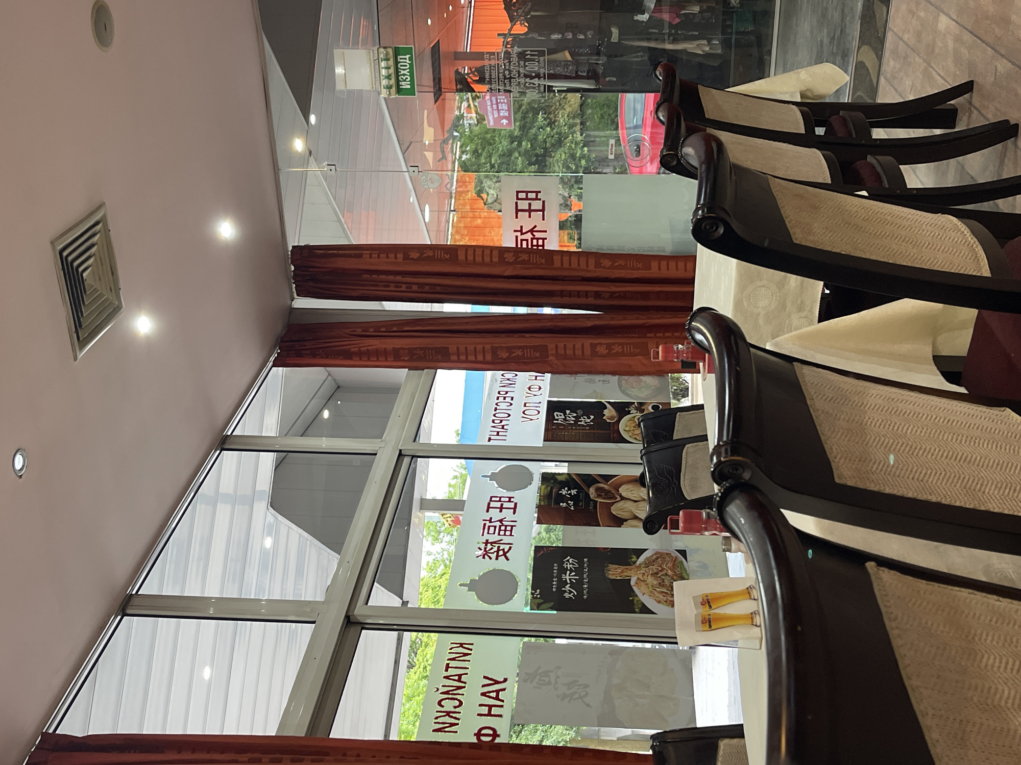 Here is a detailed description of the image:
The photograph captures the interior of a Chinese restaurant in Sofia, Bulgaria. The restaurant features large glass windows adorned with Chinese characters and advertisements for various dishes. The windows are partially covered by red curtains. The interior space is decorated with dark wooden chairs with light-colored woven seats. Tables with white tablecloths can be seen in the foreground. The sign "ИЗХОД" (EXIT) is visible in a window.